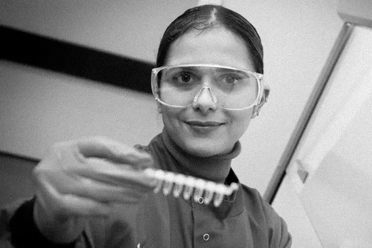 A woman in protective glasses and lab coat holding a test tube with a row of test tubes inside a laboratory.