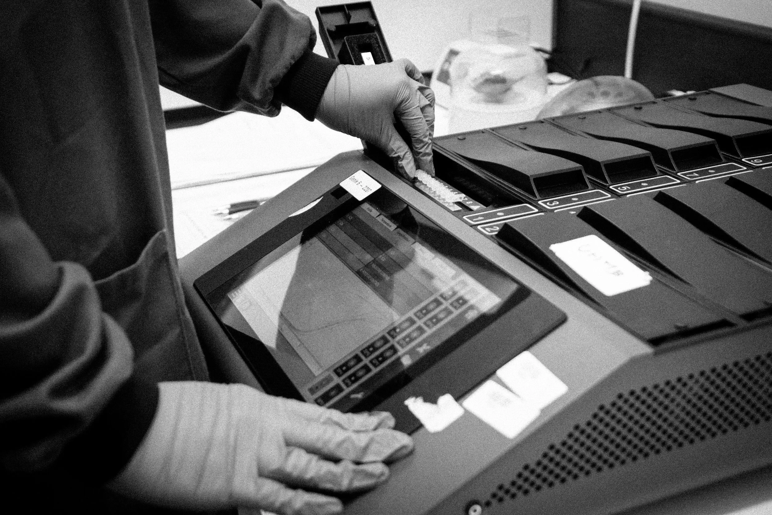 A person wearing gloves is scanning or handling medical samples or documents with a tablet device and a barcode scanner at a work station.