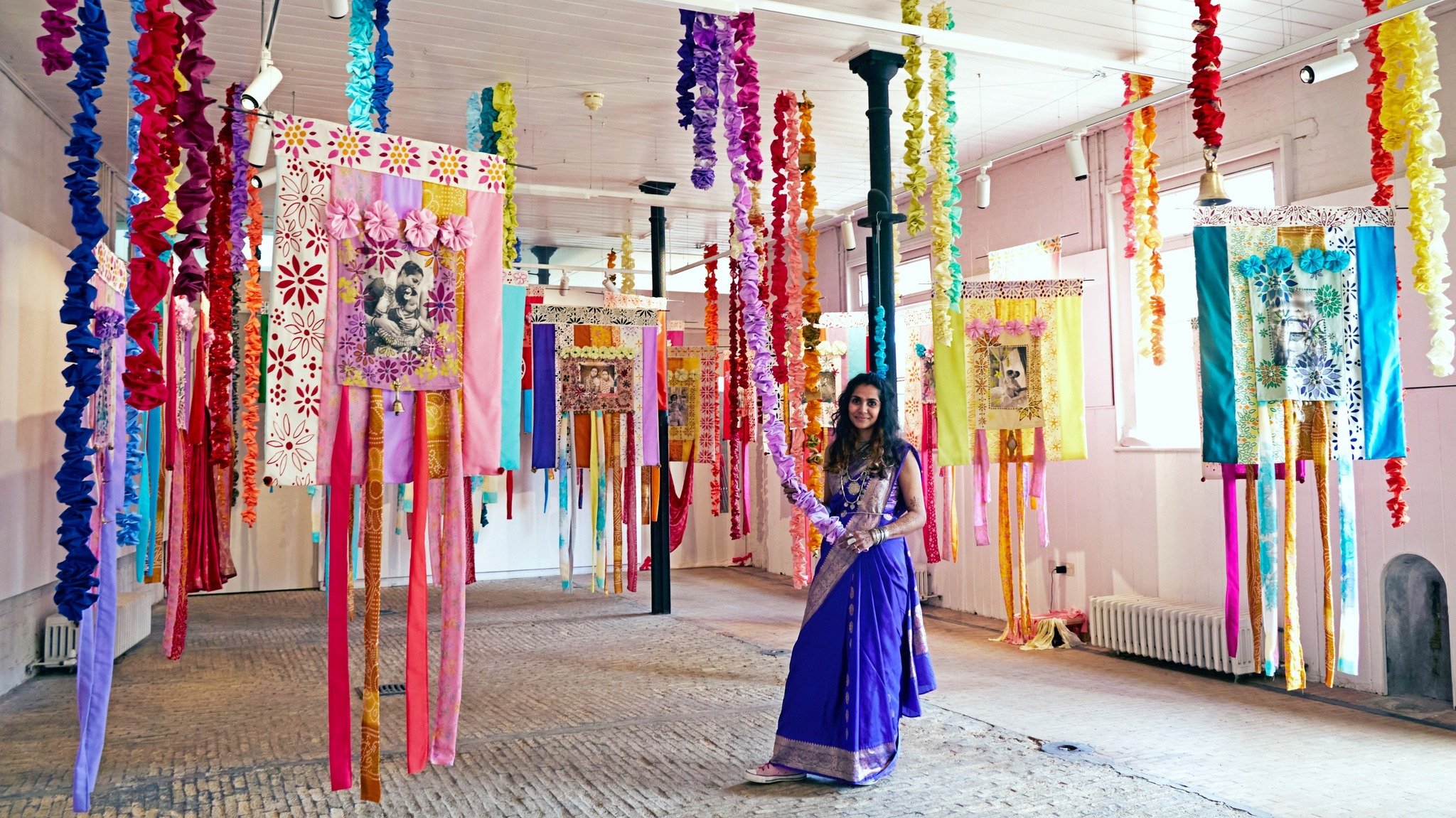 A woman in a royal blue traditional dress standing among colorful fabric banners and paper decorations hanging from the ceiling in a decorated indoor space.