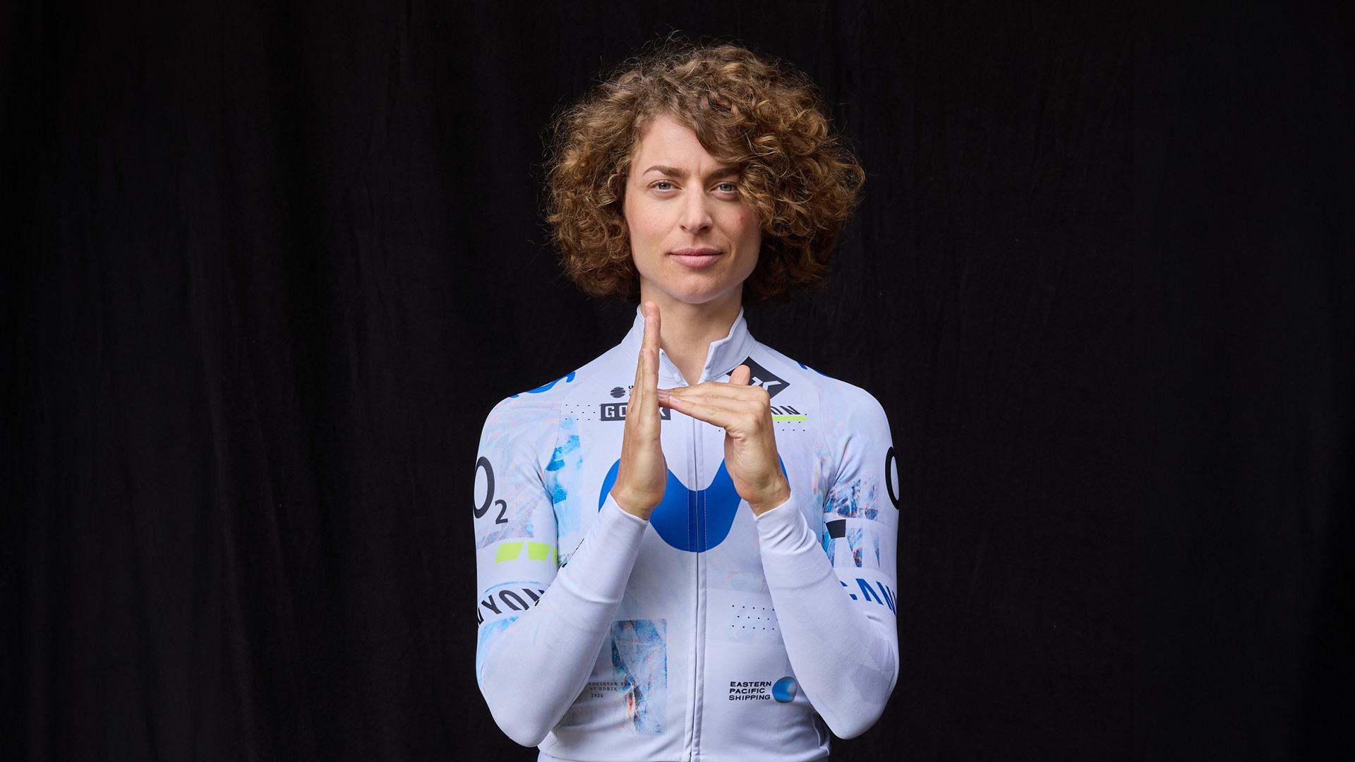 Eine Frau mit lockigem, braunem Haar in einem weißen Sporttrikot vor einem schwarzen Hintergrund, macht eine Handgeste, die wie ein Kameraobjektiv aussieht.