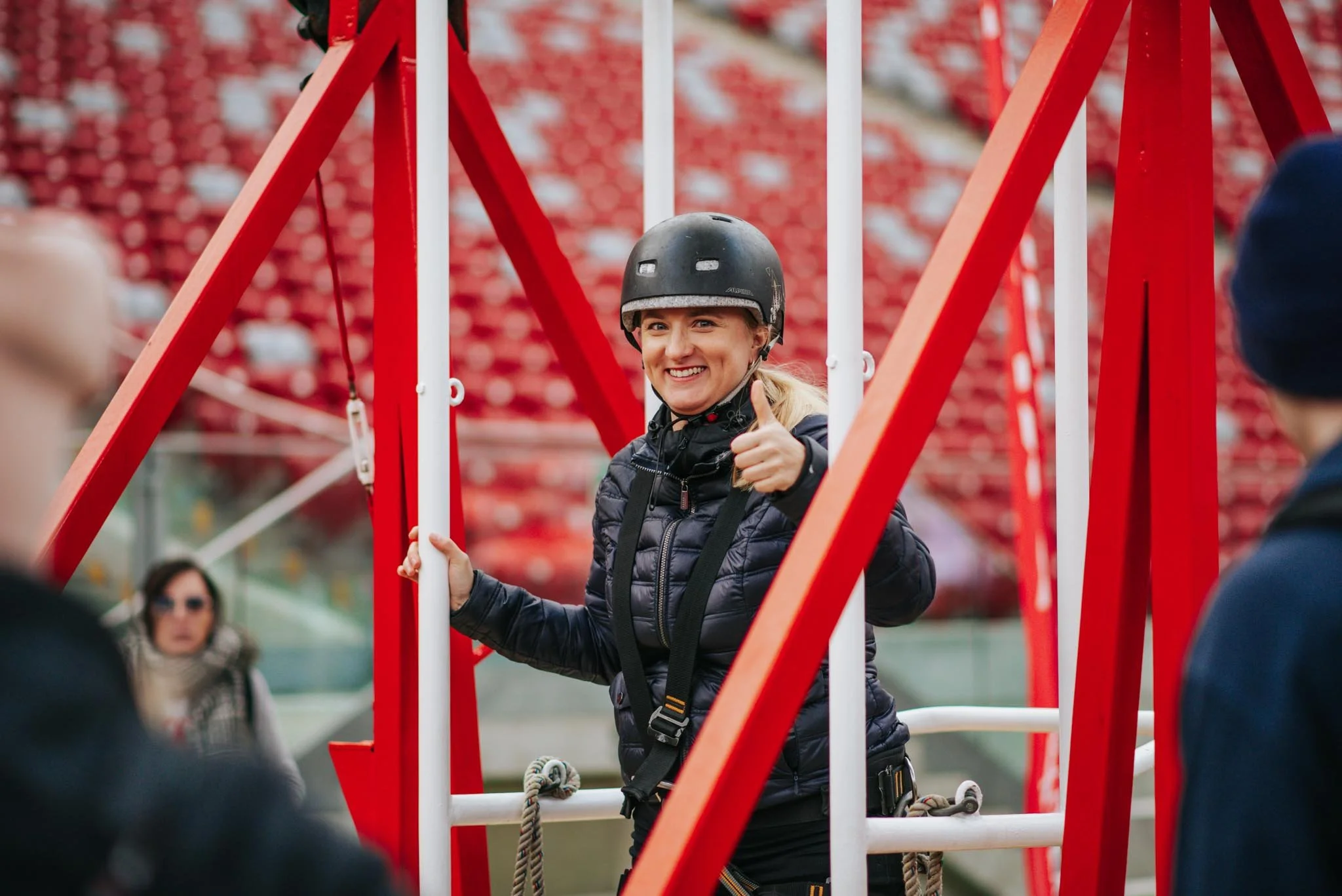 A woman wearing a helmet smiling and giving a thumbs up while standing on a swing at an outdoor venue, with red and white structures and blurred background.