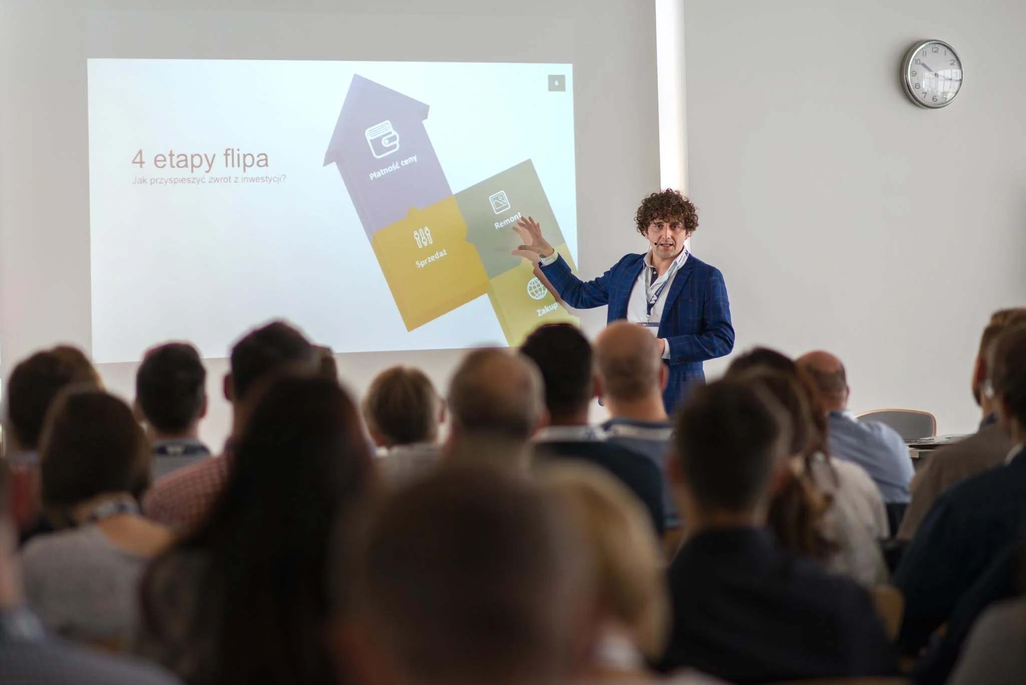 Man in blue plaid jacket giving a presentation at a conference to an audience, with a slide projected that reads "4 etapy flipa" and contains icons and text about investment steps.