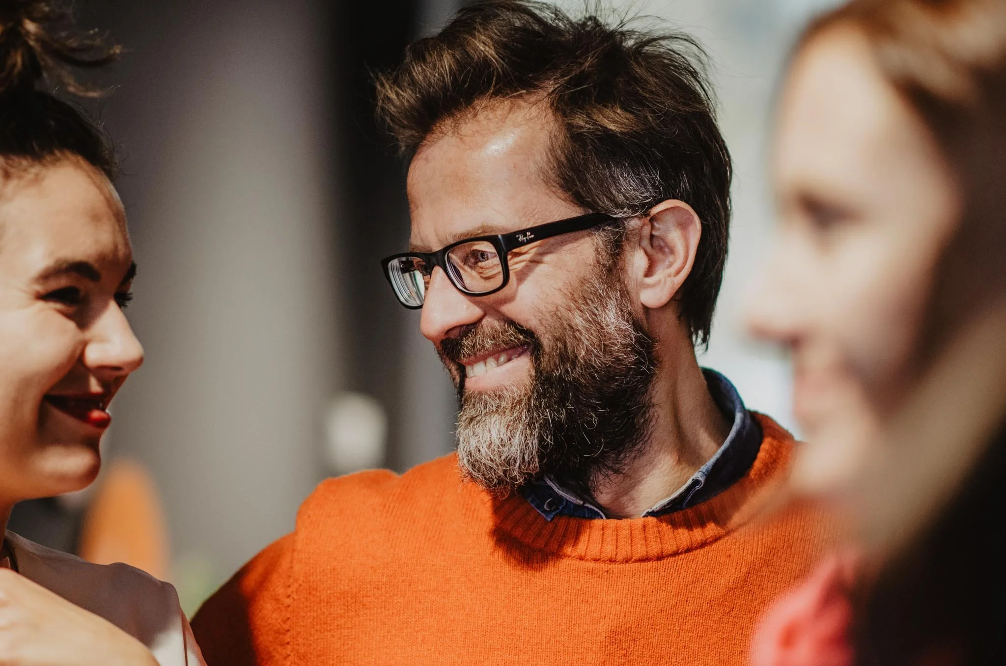 A man with glasses, a beard, and dark hair smiling and engaging with two women, one on each side, in a social setting.