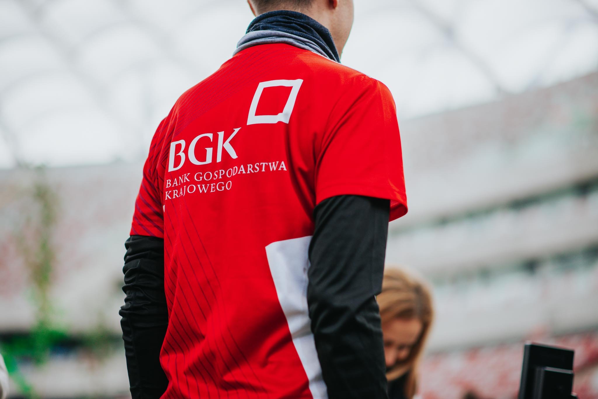Person wearing a red sports shirt with the logo 'BGK' and Polish text on the back, with a dark neck gaiter, in an indoor setting.