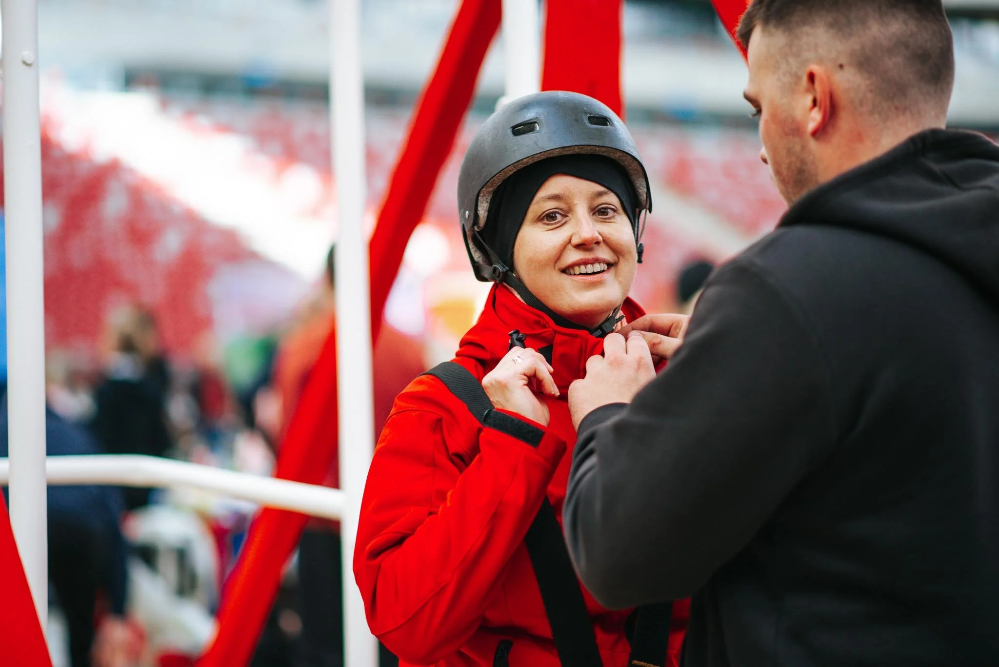 A woman wearing a helmet and red jacket smiling at a man in a black jacket at an outdoor event.