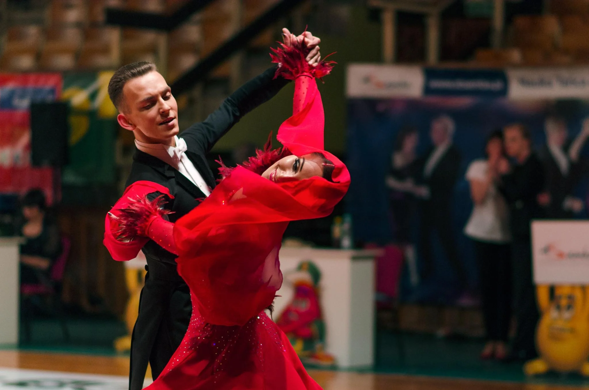 A man and woman are dancing together in a ballroom, with the man in a black tuxedo and the woman in a red dress with feathered details, during a dance competition.