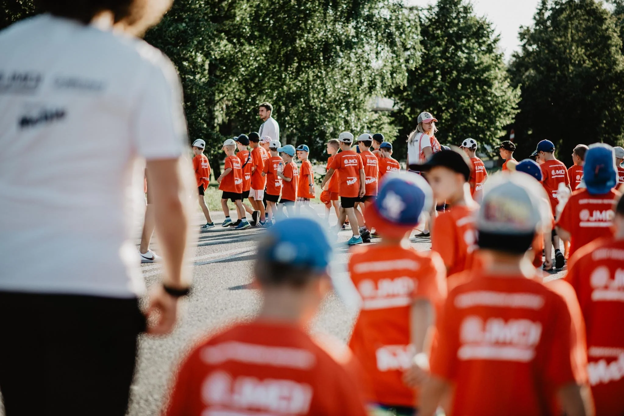 Children wearing orange shirts and caps, gathered outdoors for an event or activity. A man with a beard stands in the background among trees.