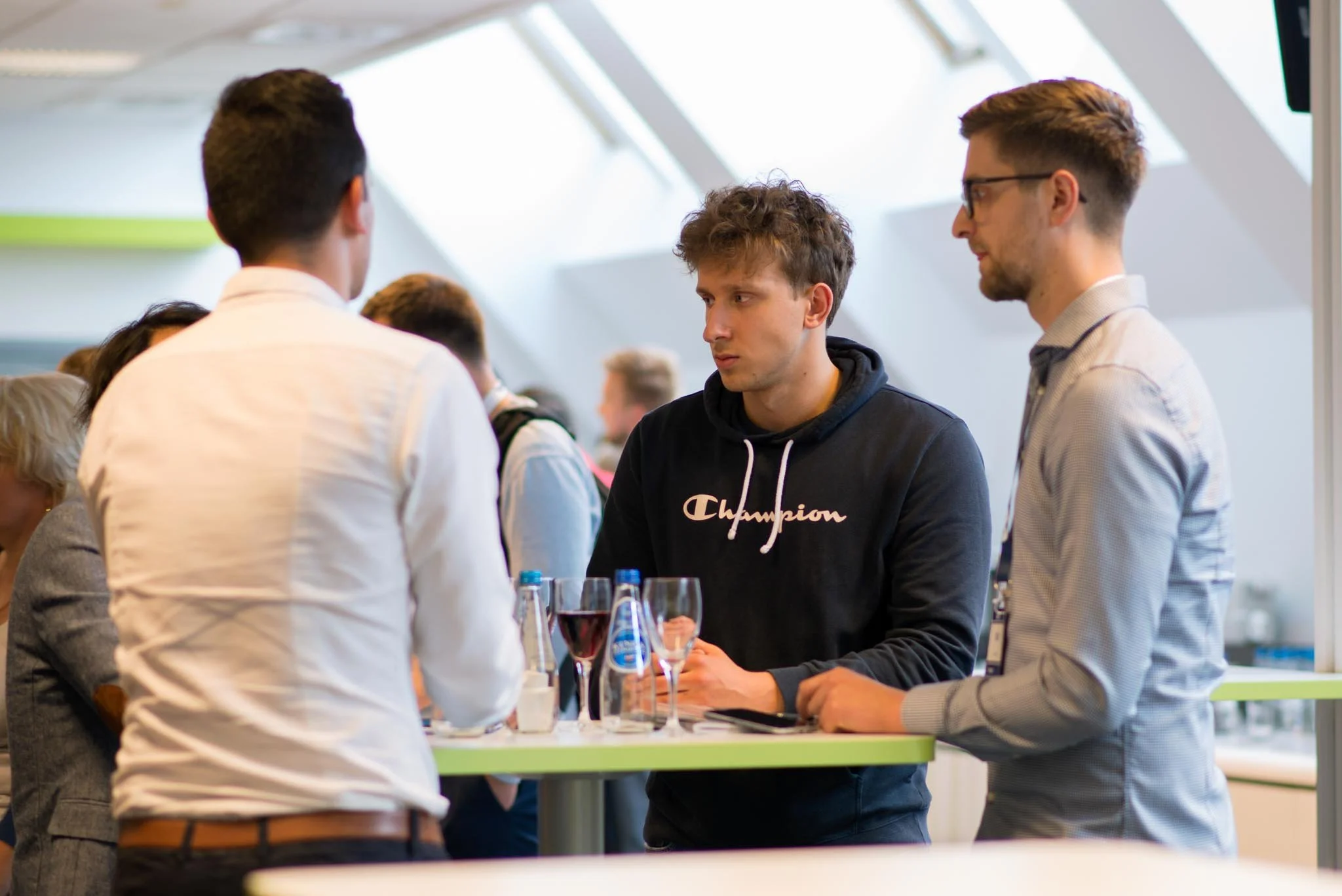 Three young men having conversation at a social event, with a table of drinks in front of them, indoors with a modern ceiling design.