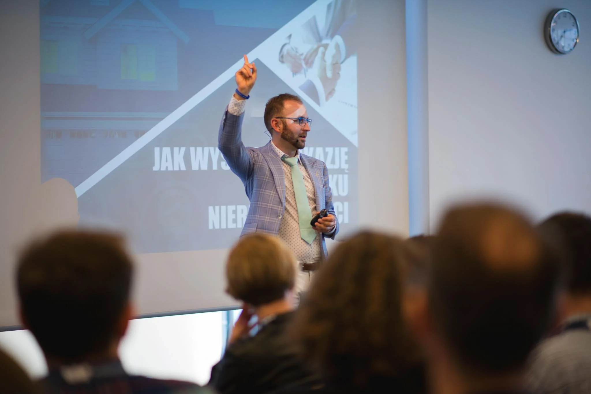 Man giving a presentation in front of an audience, pointing upward, with a projector screen displaying a slide behind him.