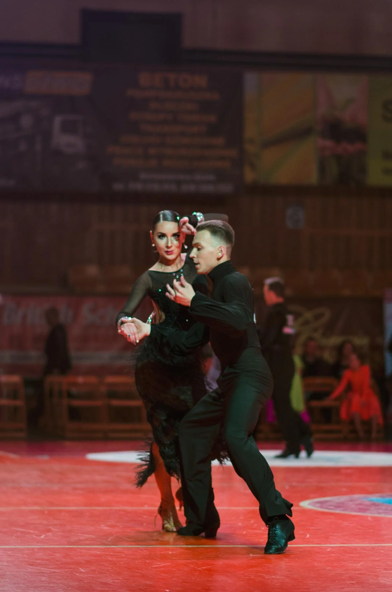 A couple performing a ballroom dance in a dance competition, with a woman in a black dress with feathers and a man in black attire, dancing on a red floor with a blurred background.
