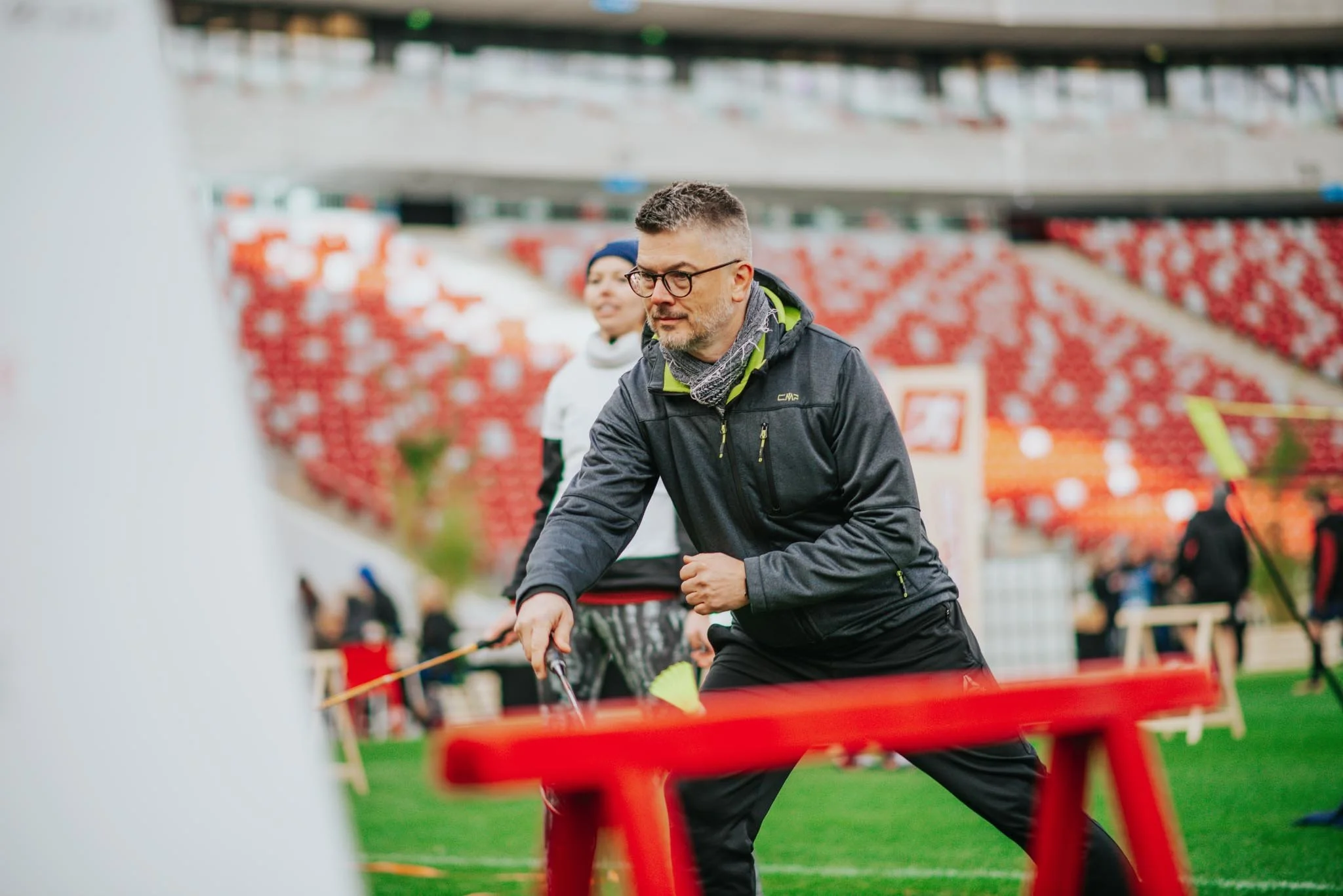Two people, a man and a woman, working outdoors in a stadium with red seat rows, engaged in planting or gardening activities, with green grass and trees in the background.
