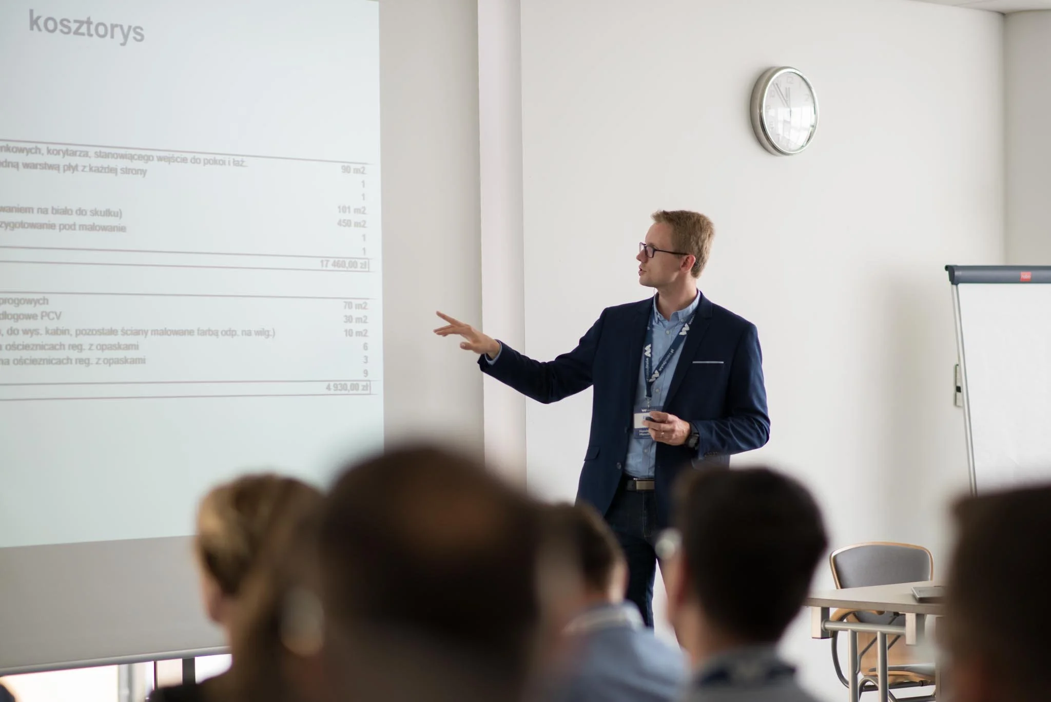 A man in a suit and glasses presents to a group of people in a conference room, with a slide projected on the wall behind him containing text in Polish and a clock on the wall showing nearly 12:05.