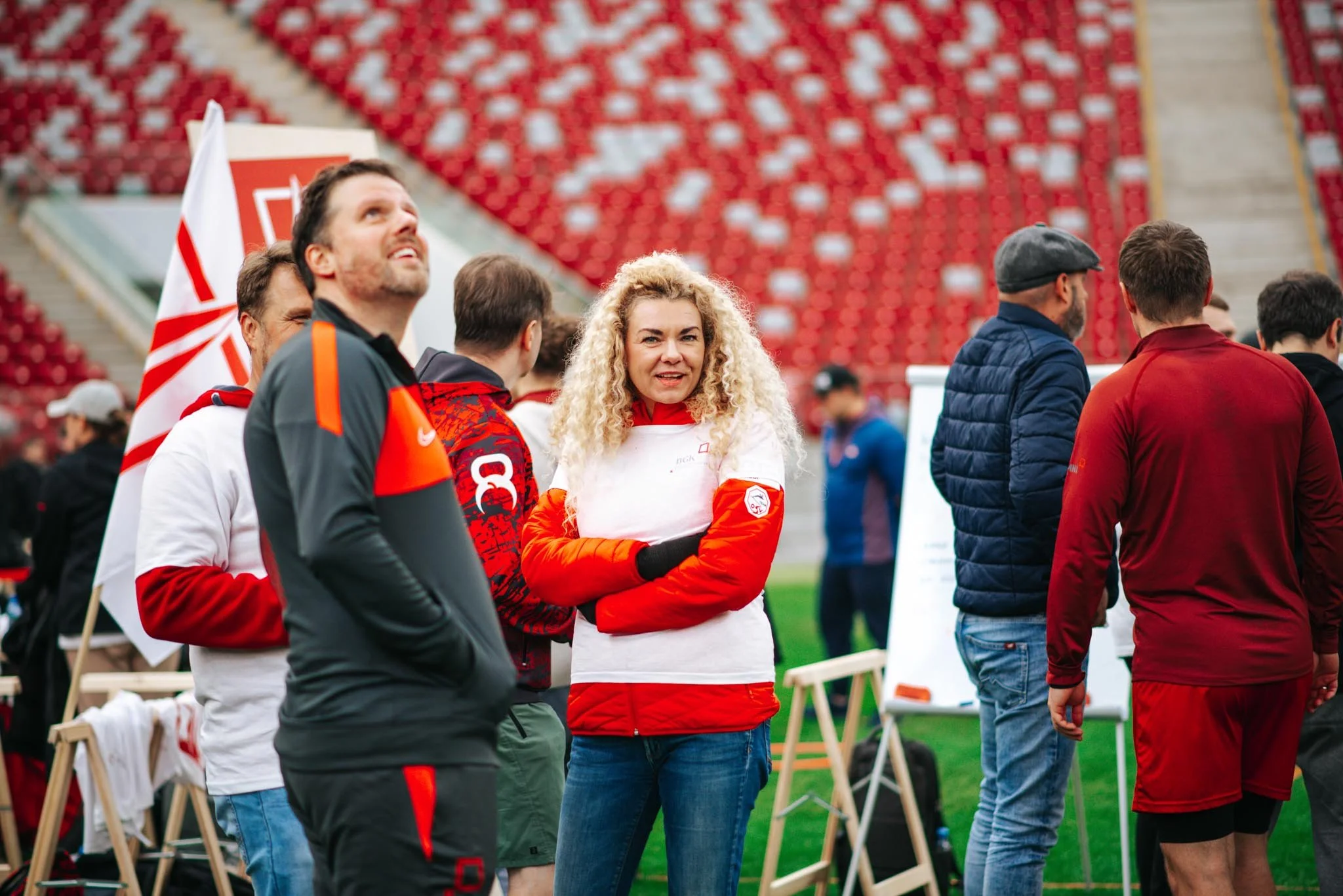 A group of people, including a woman with curly blonde hair and others wearing sports jerseys, gathered at a sports stadium. They appear to be socializing or preparing for an event, with red stadium seats and a field in the background.