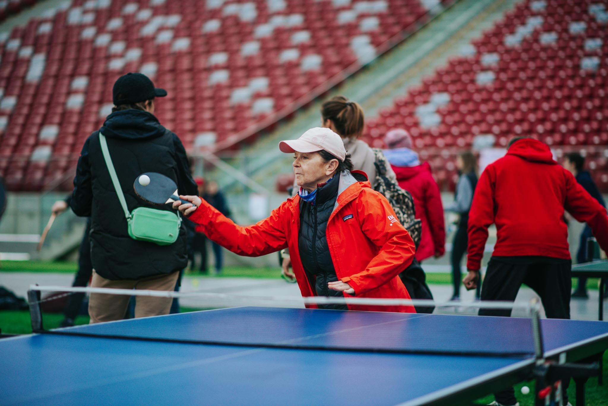 People playing ping pong outdoors at a park, with a woman in a red jacket and white cap in the foreground, and others in the background near a large red and beige outdoor stadium seating.