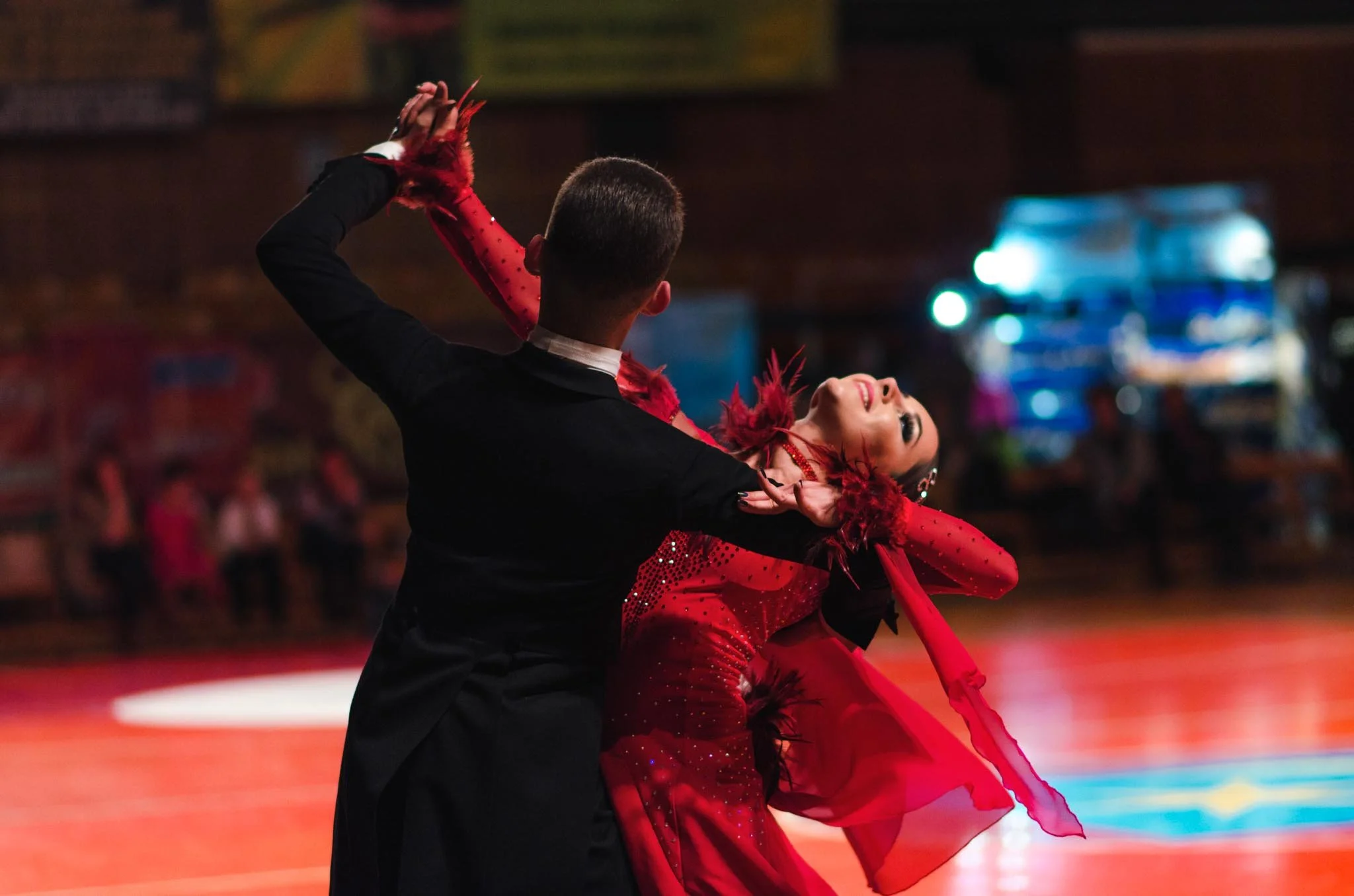 A couple ballroom dancing on a brightly lit dance floor, with the woman wearing a red dress with feathered accents and the man in a black tuxedo.