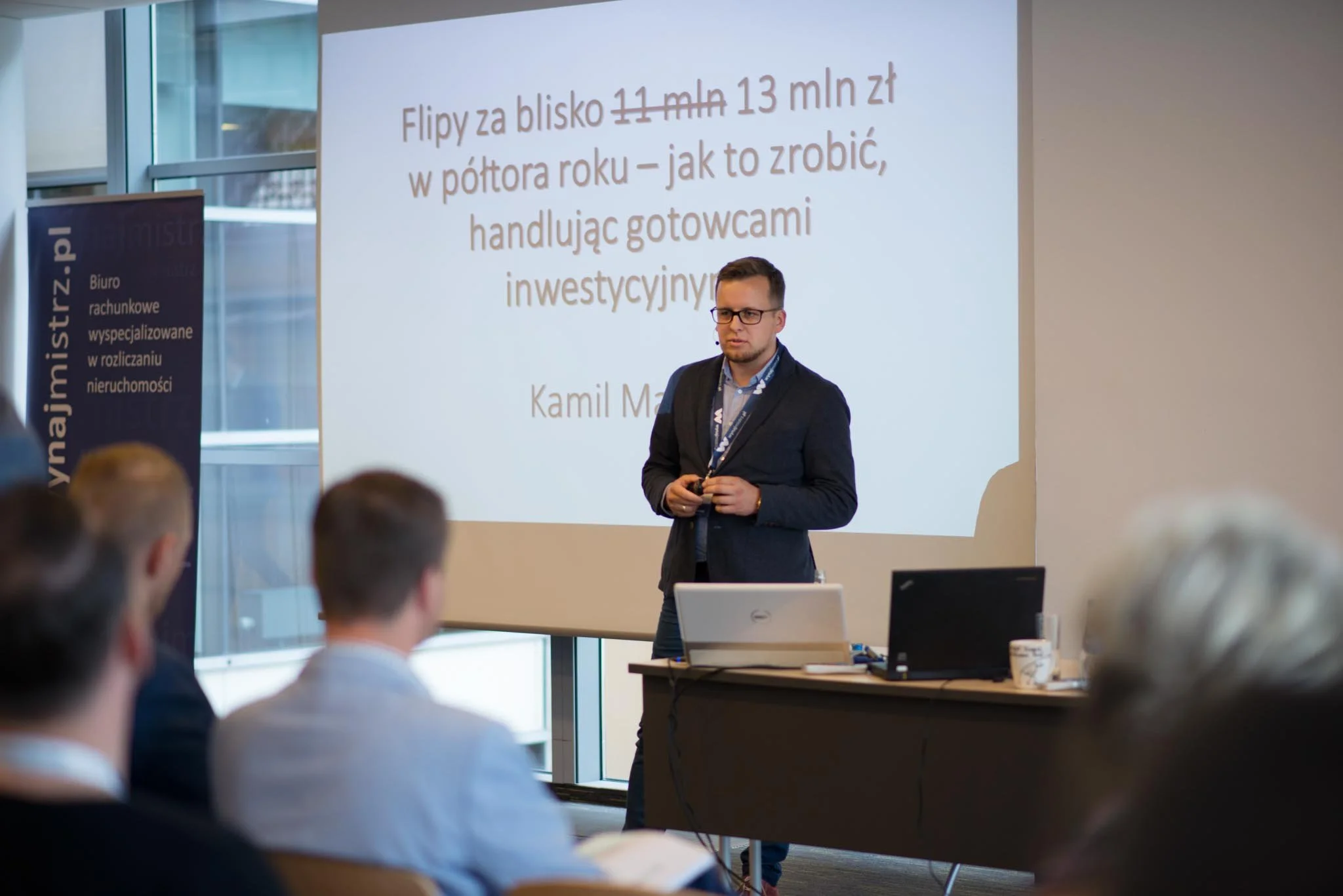 A man in glasses and a dark jacket standing in front of a large presentation screen with Polish text, speaking to an audience in a conference room.