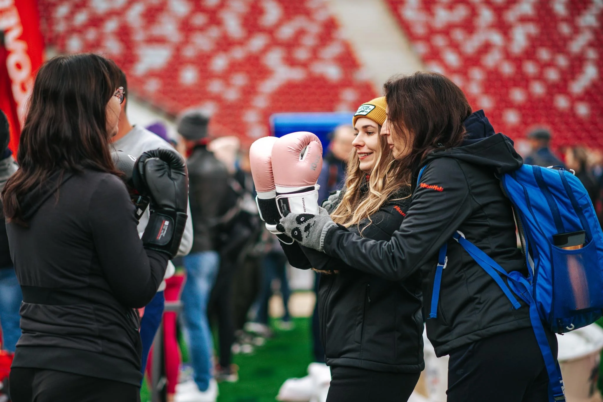 Two women in jackets and gloves exchanging boxing gloves during an outdoor fitness event, with other people and a red patterned background in the scene.