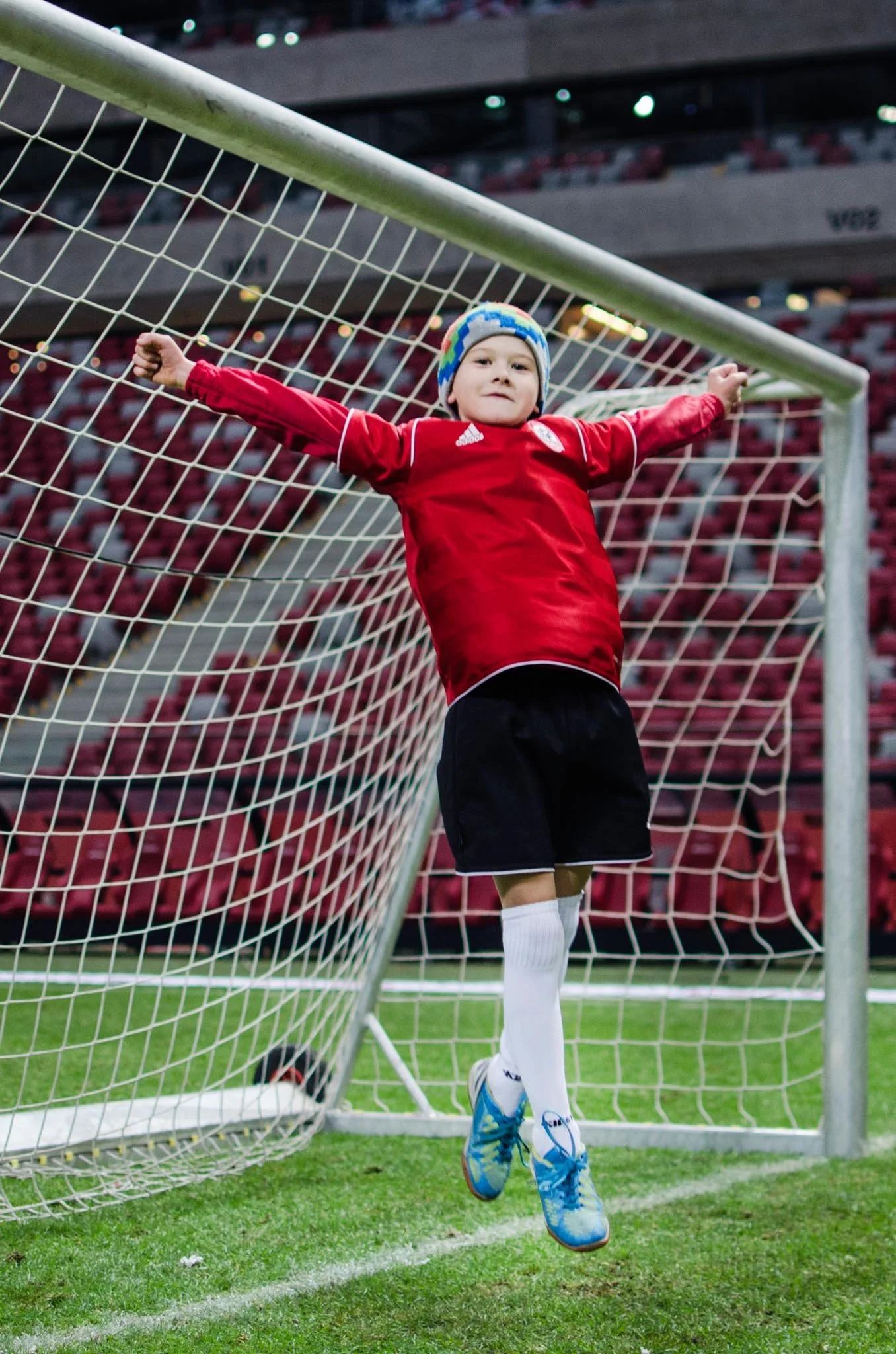 A young boy in a red soccer jersey, shorts, and blue soccer shoes is standing inside the goalpost on a soccer field, with his arms outstretched and leaning against the net, in a stadium with red seating.