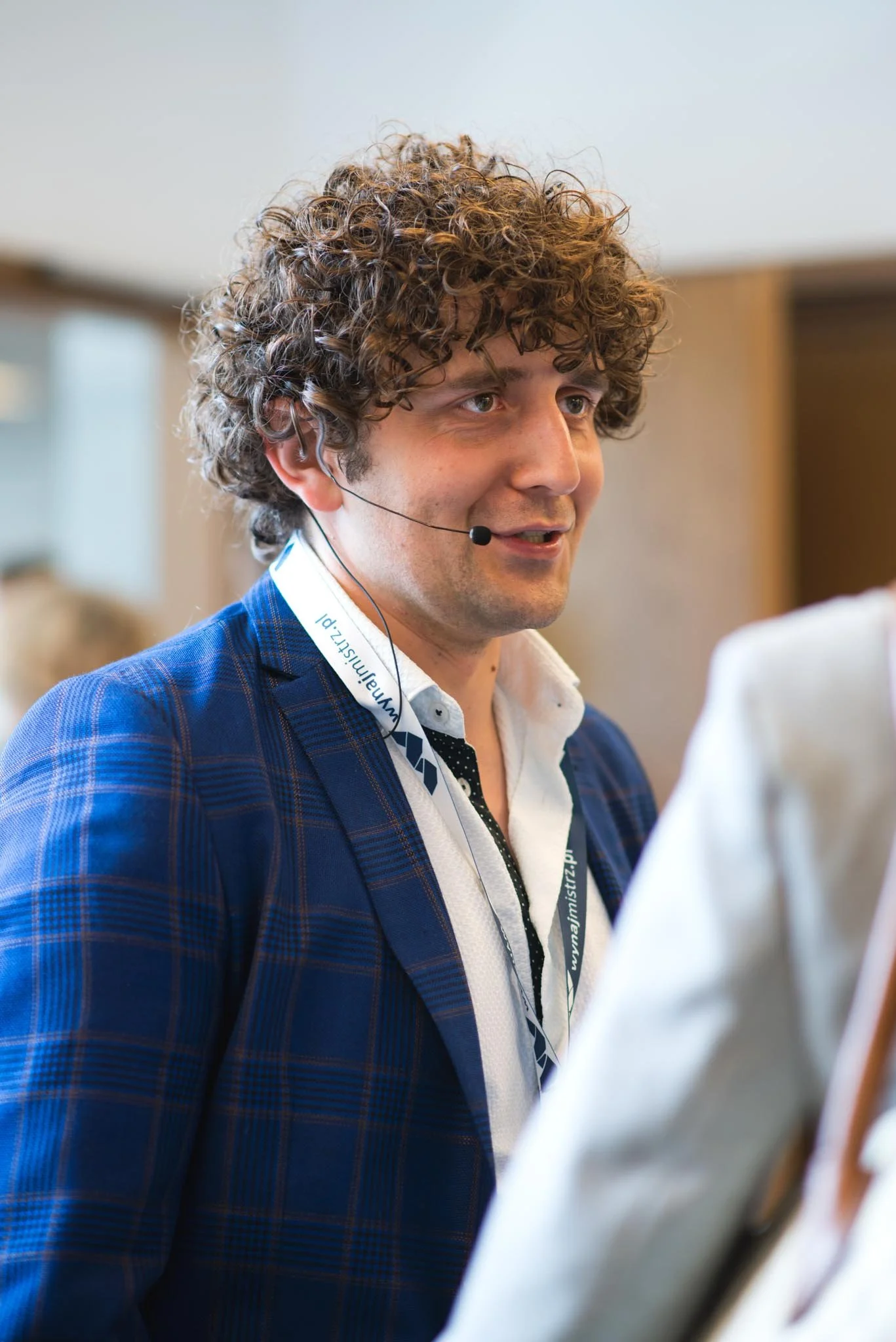 A man with curly brown hair and a headset, wearing a blue plaid suit jacket and a white shirt, speaking during a presentation or conference.
