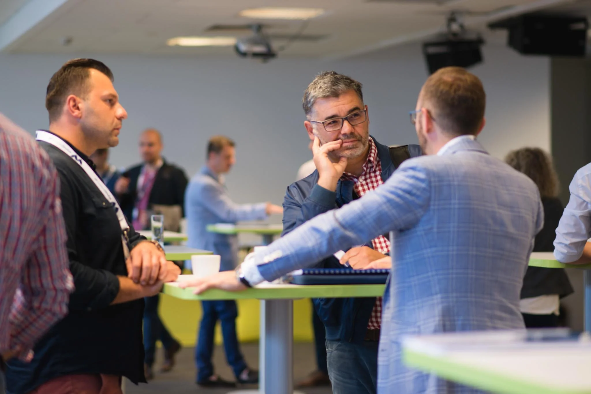 Three men, two of whom are wearing glasses, are engaged in conversation in a modern conference room with high-top tables. The middle man is touching his face while listening. There are other people blurred in the background.