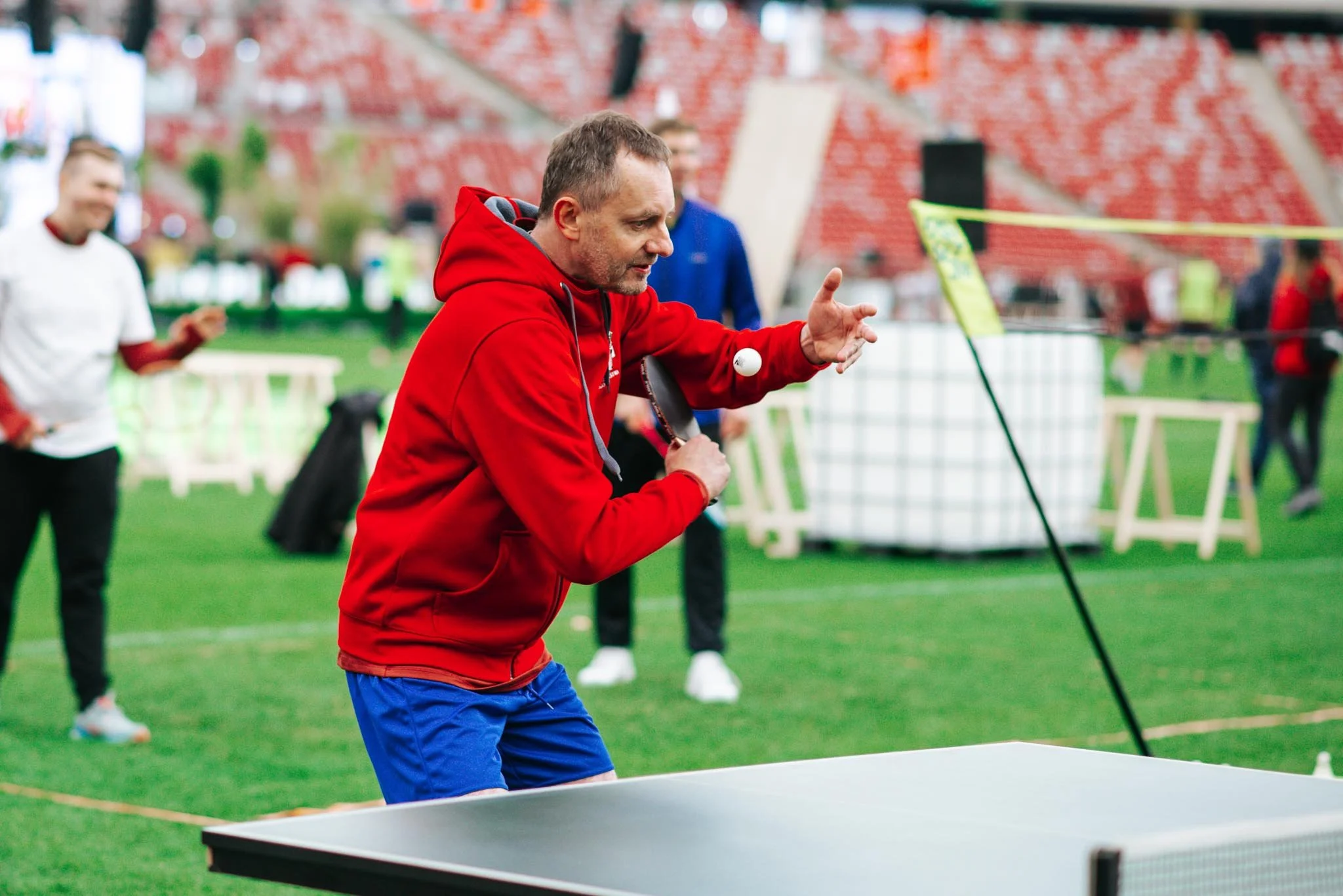 A man in a red hoodie and blue shorts playing ping pong in an indoor sports facility with a green floor. Other people are visible in the background, some playing and some watching.