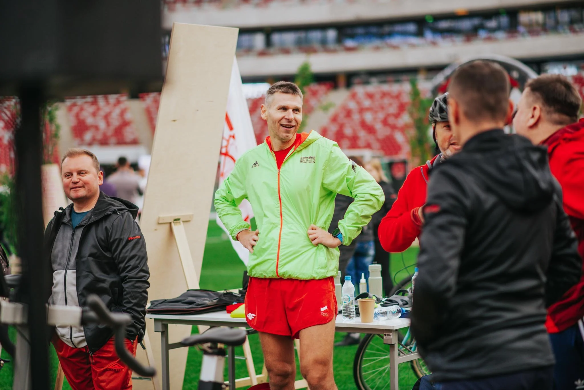 A male athlete in red shorts and a bright green jacket standing and talking to a group of people at an outdoor event in a stadium.