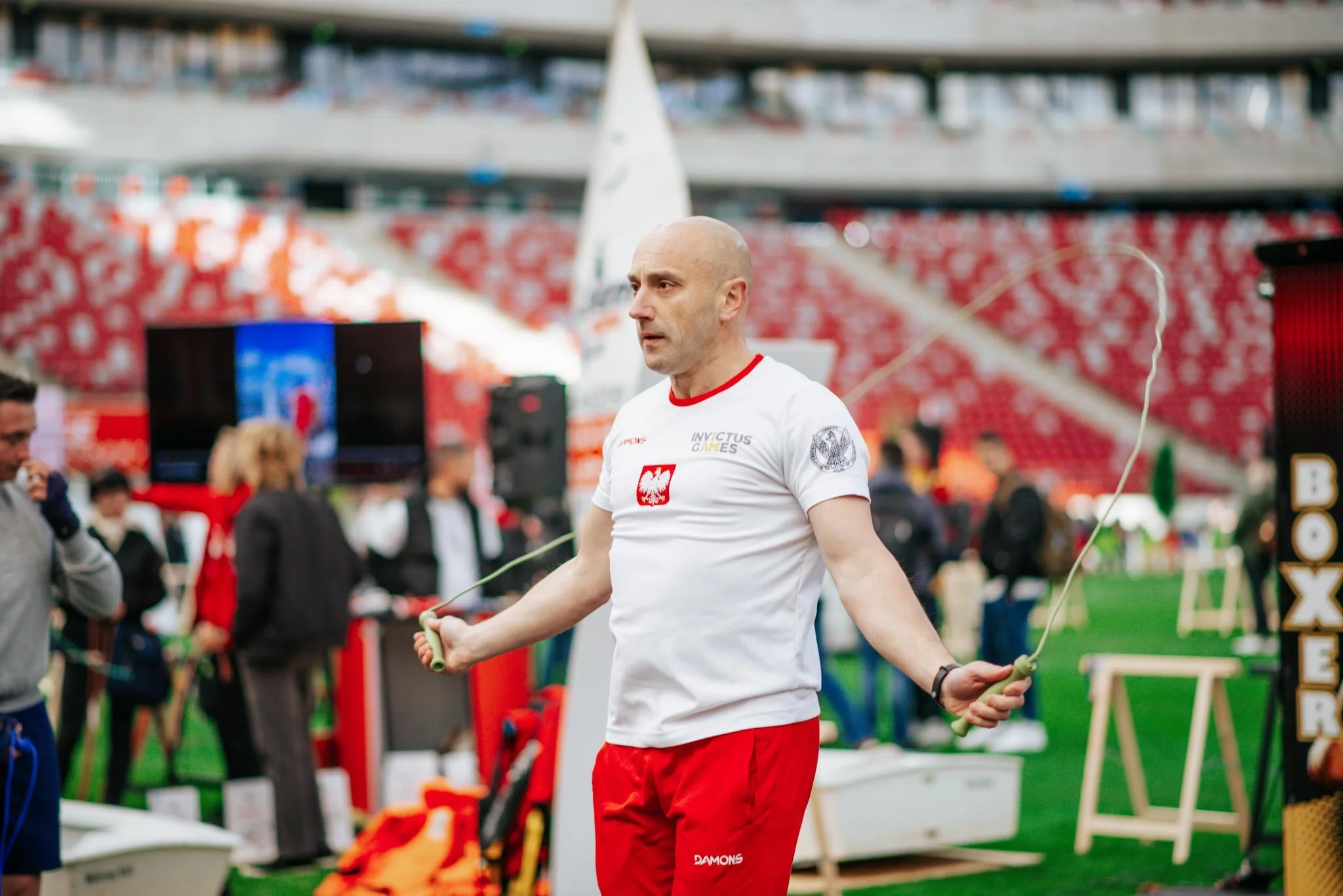A man with a shaved head wearing a white t-shirt with a red emblem and red shorts holding a jump rope in an athletic stadium. There are people in the background along with various equipment and banners.
