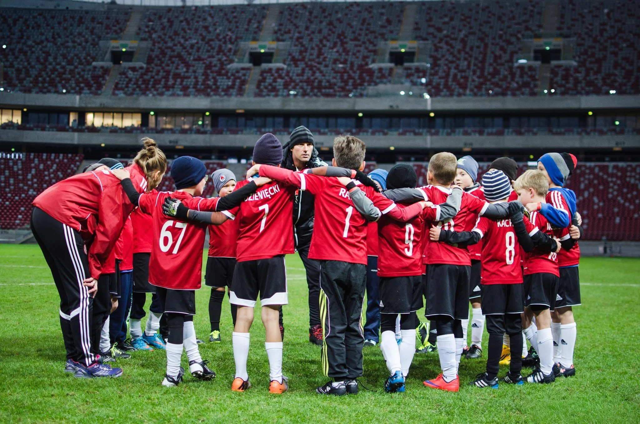 Young soccer players in red uniforms and black shorts huddle with their coach on a soccer field in a large stadium, preparing for a game or practice.