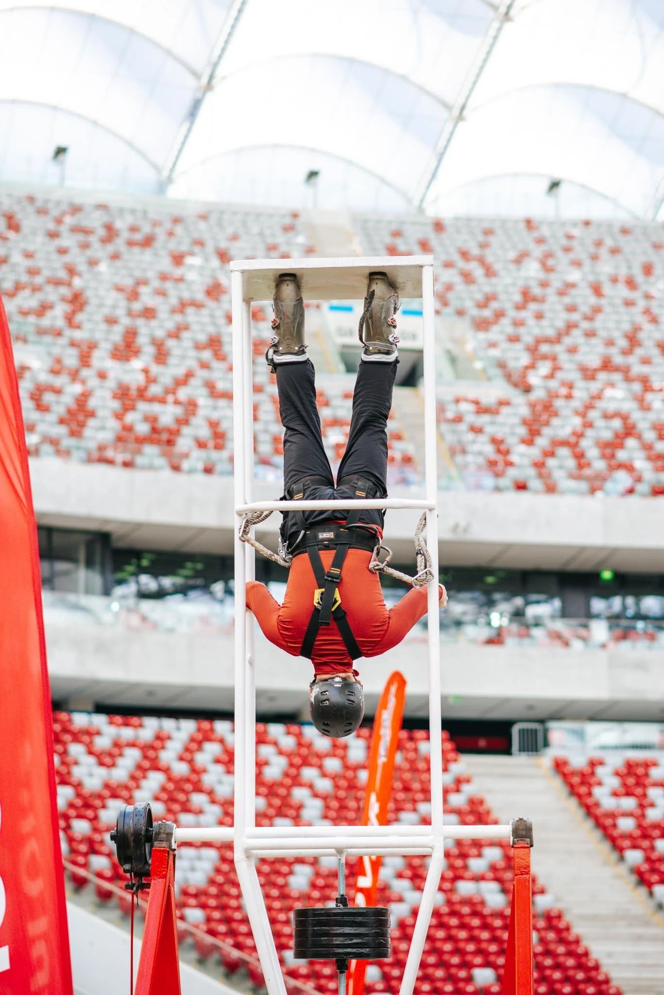 A person in a red jacket and black helmet performing an upside-down stunt on a white apparatus in a stadium.