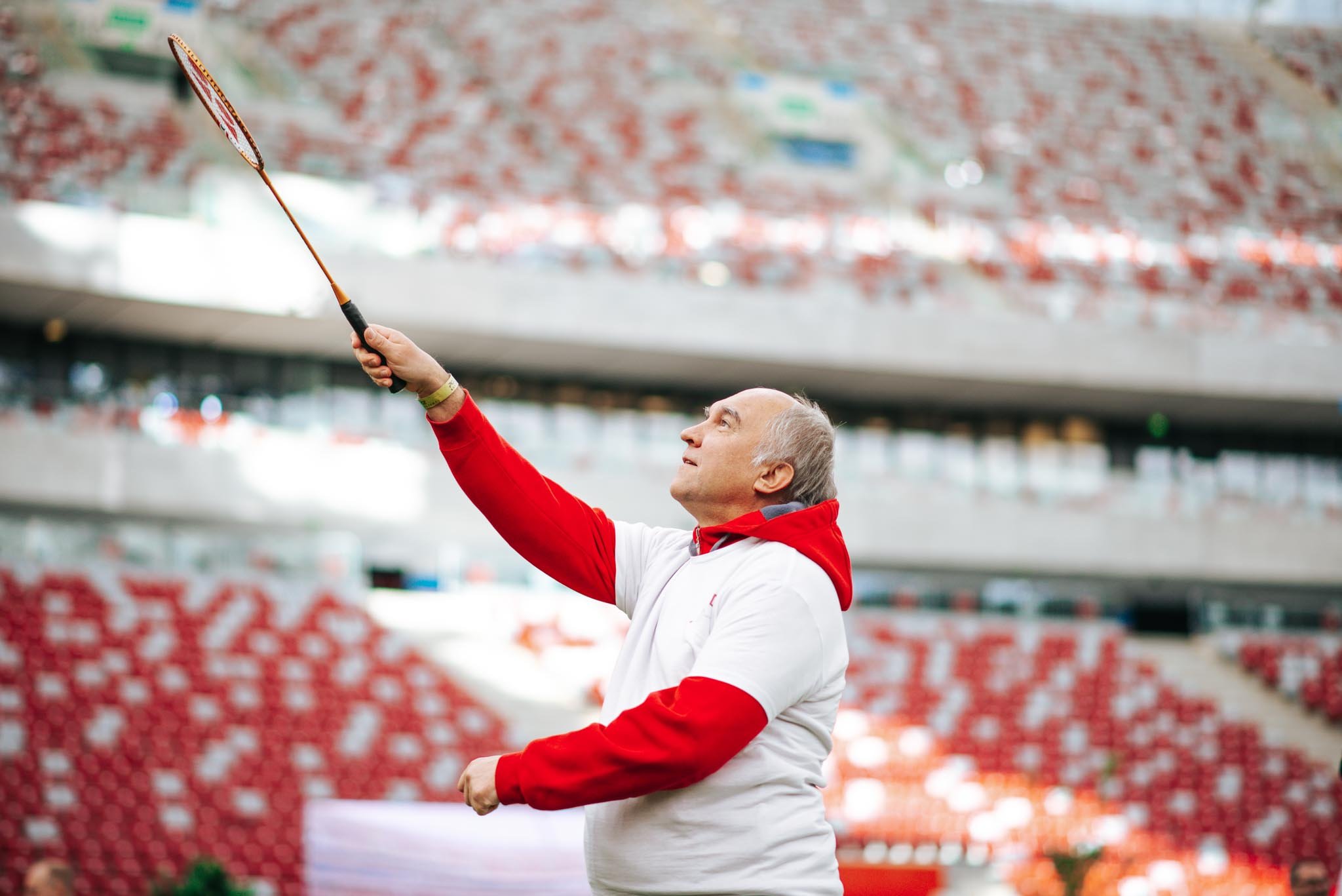 A man with short gray hair wearing a red and white jacket is holding a badminton racket high with his right hand, looking up at it. The background shows a blurred stadium with red seating.