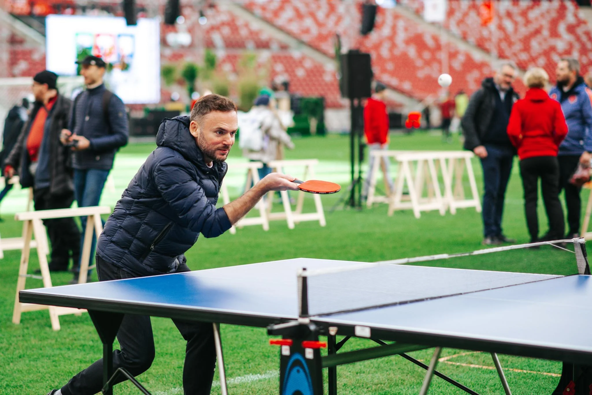 A man in a blue jacket playing table tennis indoors, with several people and a red patterned wall in the background.