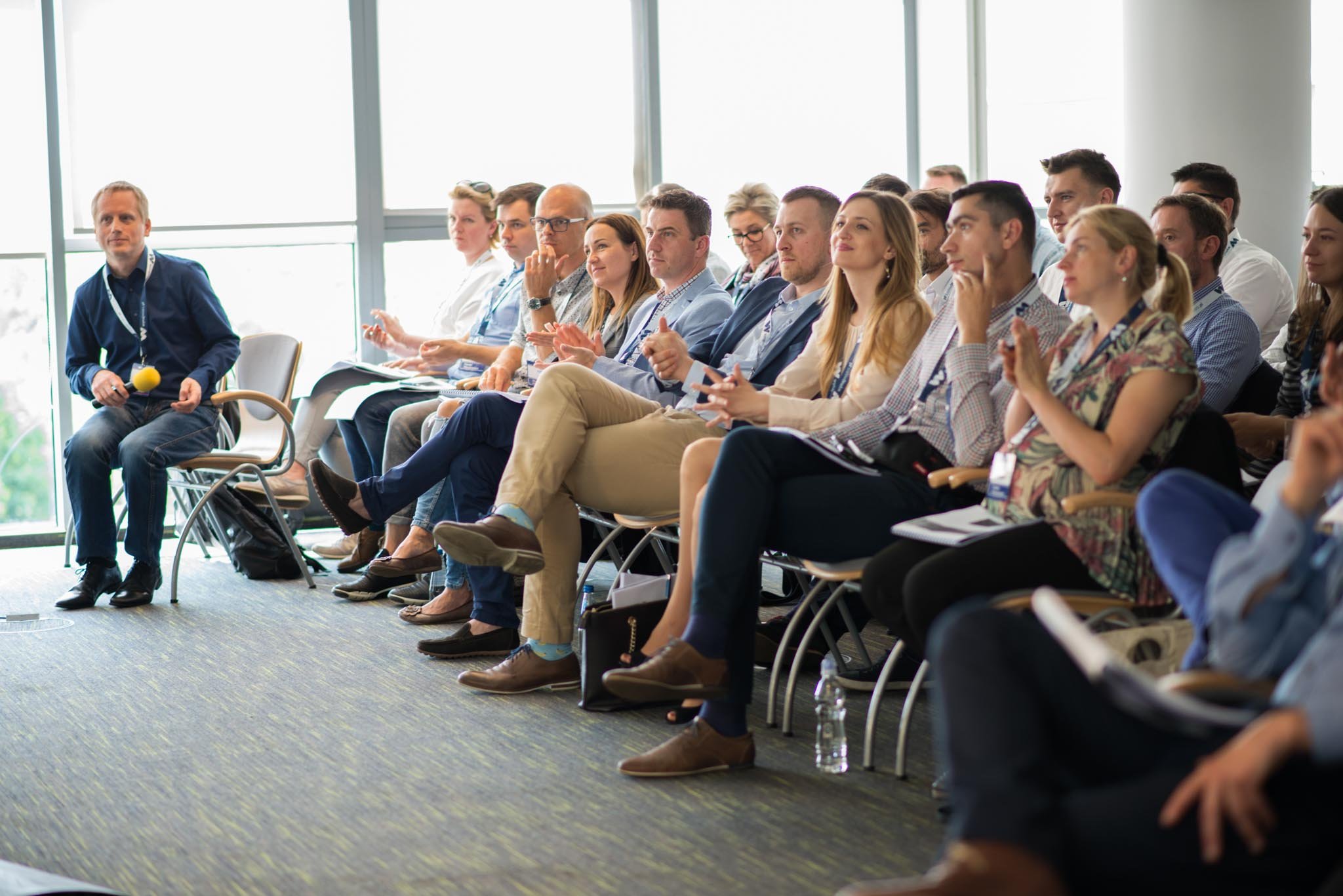 A group of people attending a conference or seminar, sitting in chairs and listening attentively, in a room with large windows.