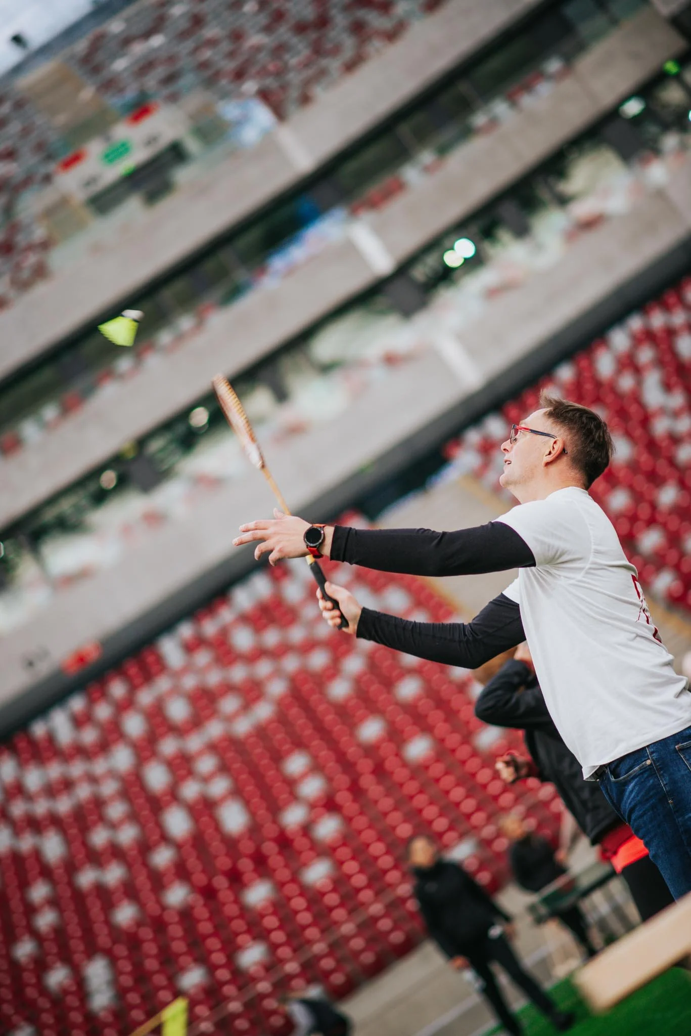 A man with glasses and a smartwatch playing badminton outdoors, with a red stadium seating background.