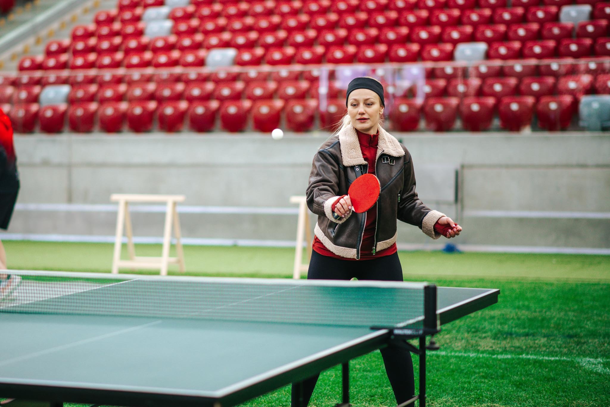 A woman playing table tennis in an empty stadium, holding a red paddle and dressed warmly in a coat and headband, with rows of red stadium seats in the background.