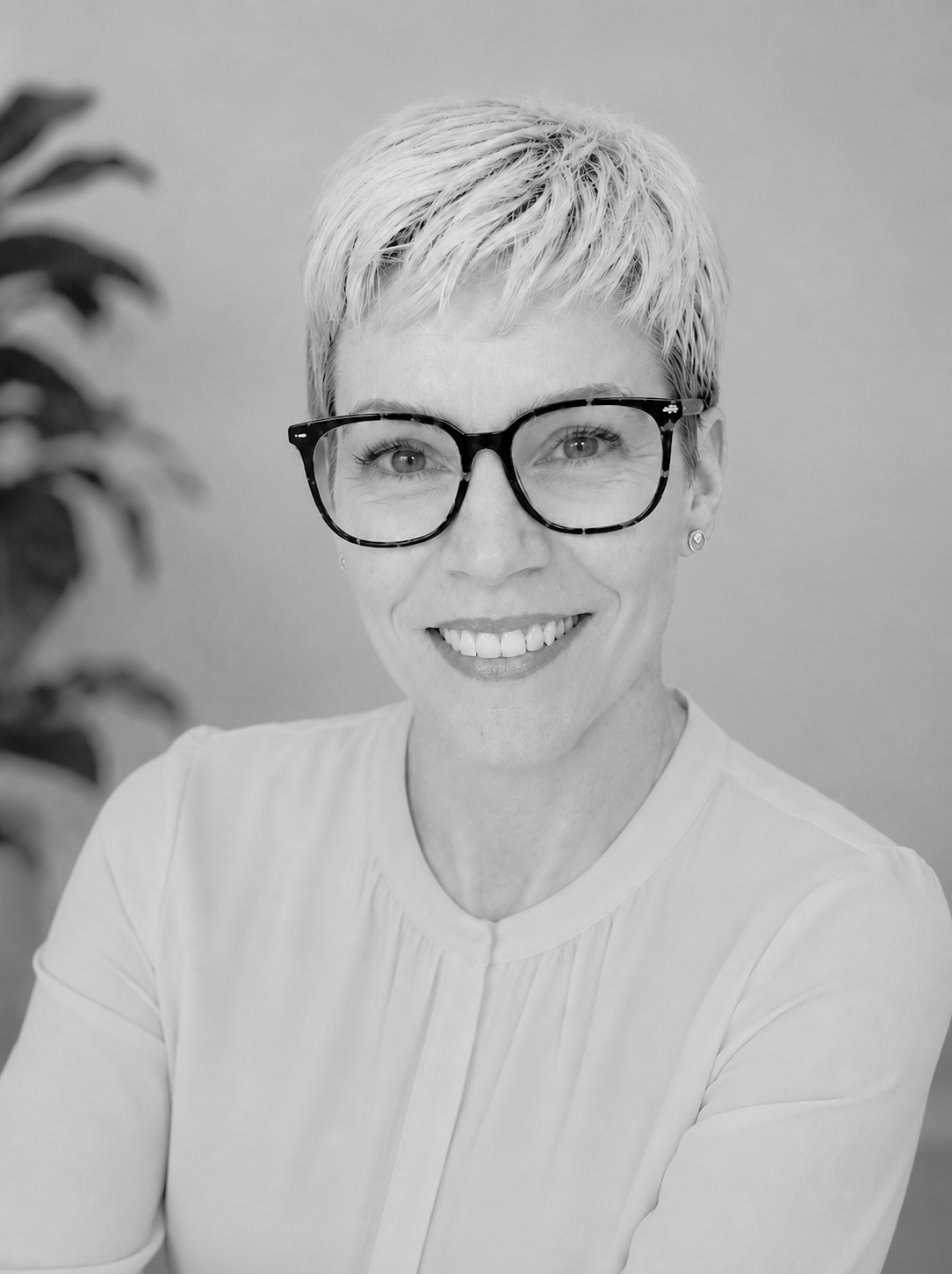 Black and white portrait of a woman with short platinum blonde hair, wearing large black glasses, smiling, in a short-sleeved top, with a plant in the background.