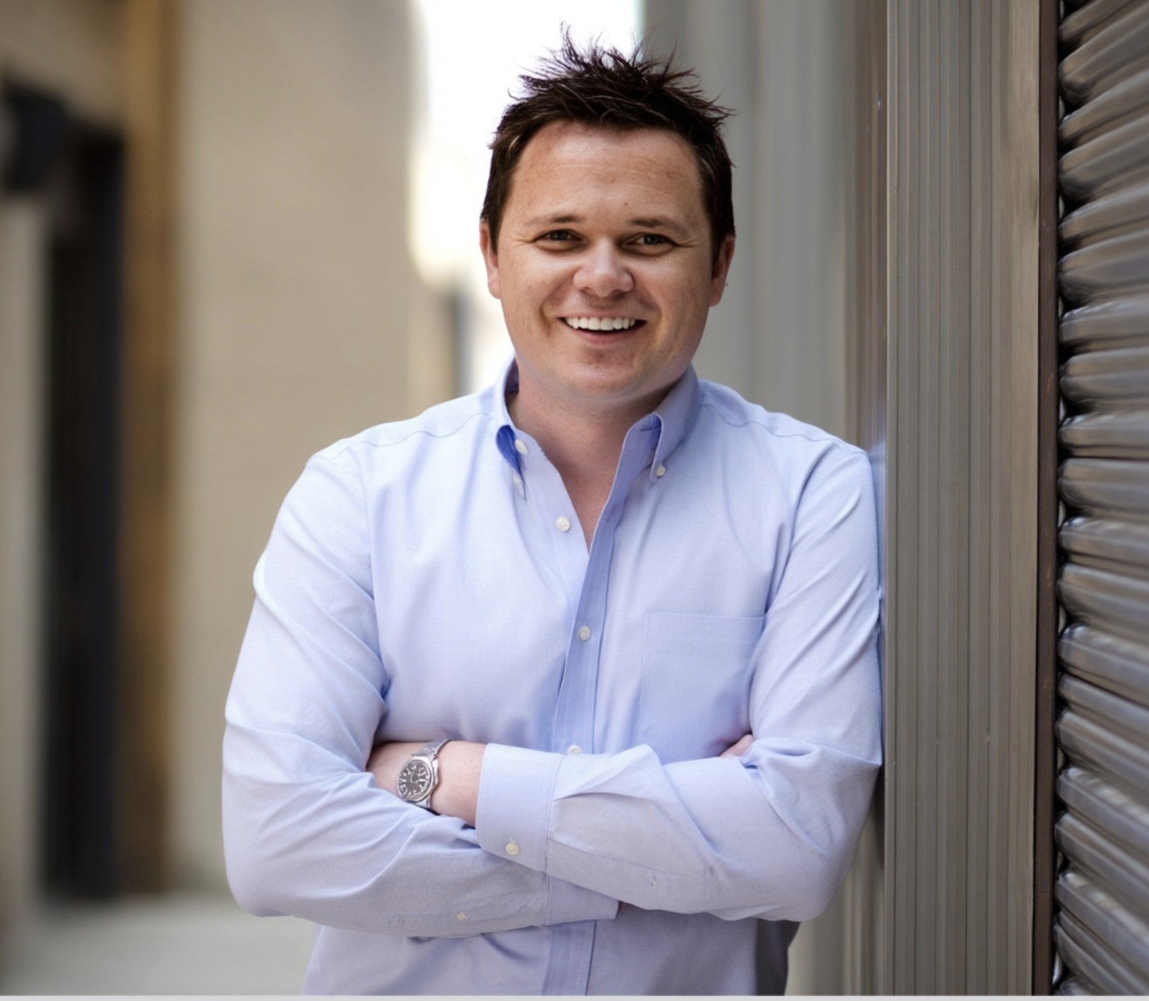 A smiling man with dark hair, wearing a light blue button-up shirt, standing outdoors between two metal shutter doors with arms crossed.