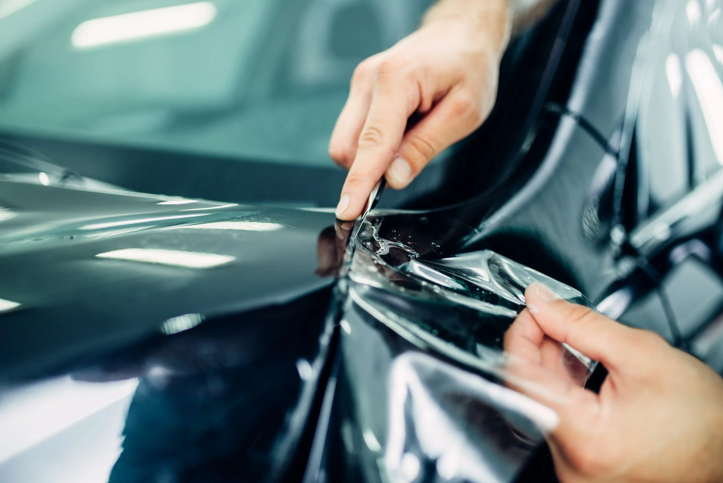 Close-up of hands applying a protective film to a dark-colored car's body.