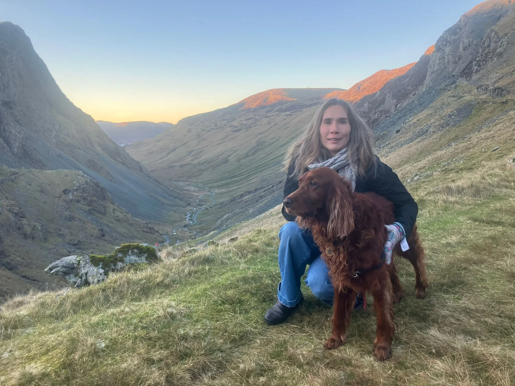 A woman with long hair in a black jacket and blue jeans sitting in a grassy valley with her dog, surrounded by mountains during sunset.