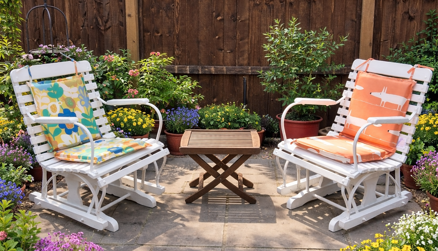 Two white outdoor patio chairs with colorful cushions facing each other, with a small wooden folding table between them, surrounded by potted flowers and plants in a backyard garden.