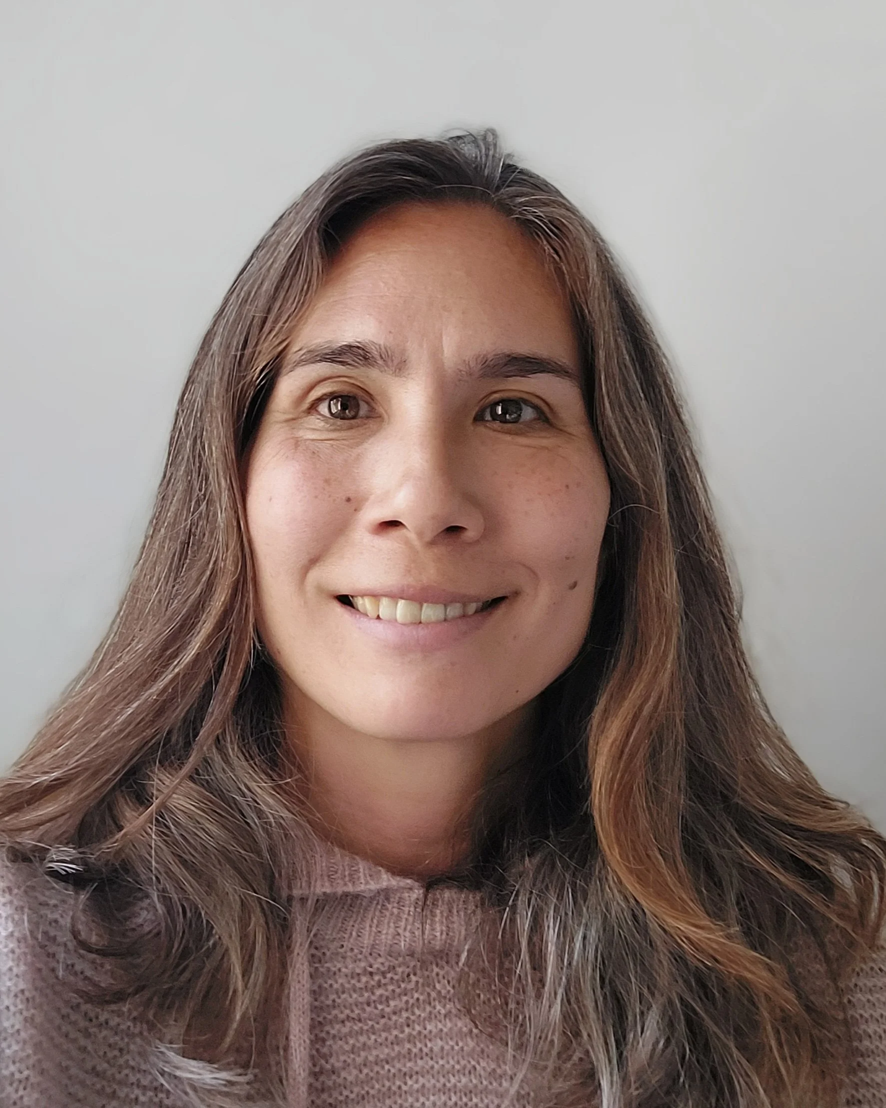 Close-up of a smiling woman with long brown hair and brown eyes, wearing a pinkish-brown sweater, against a plain white background.