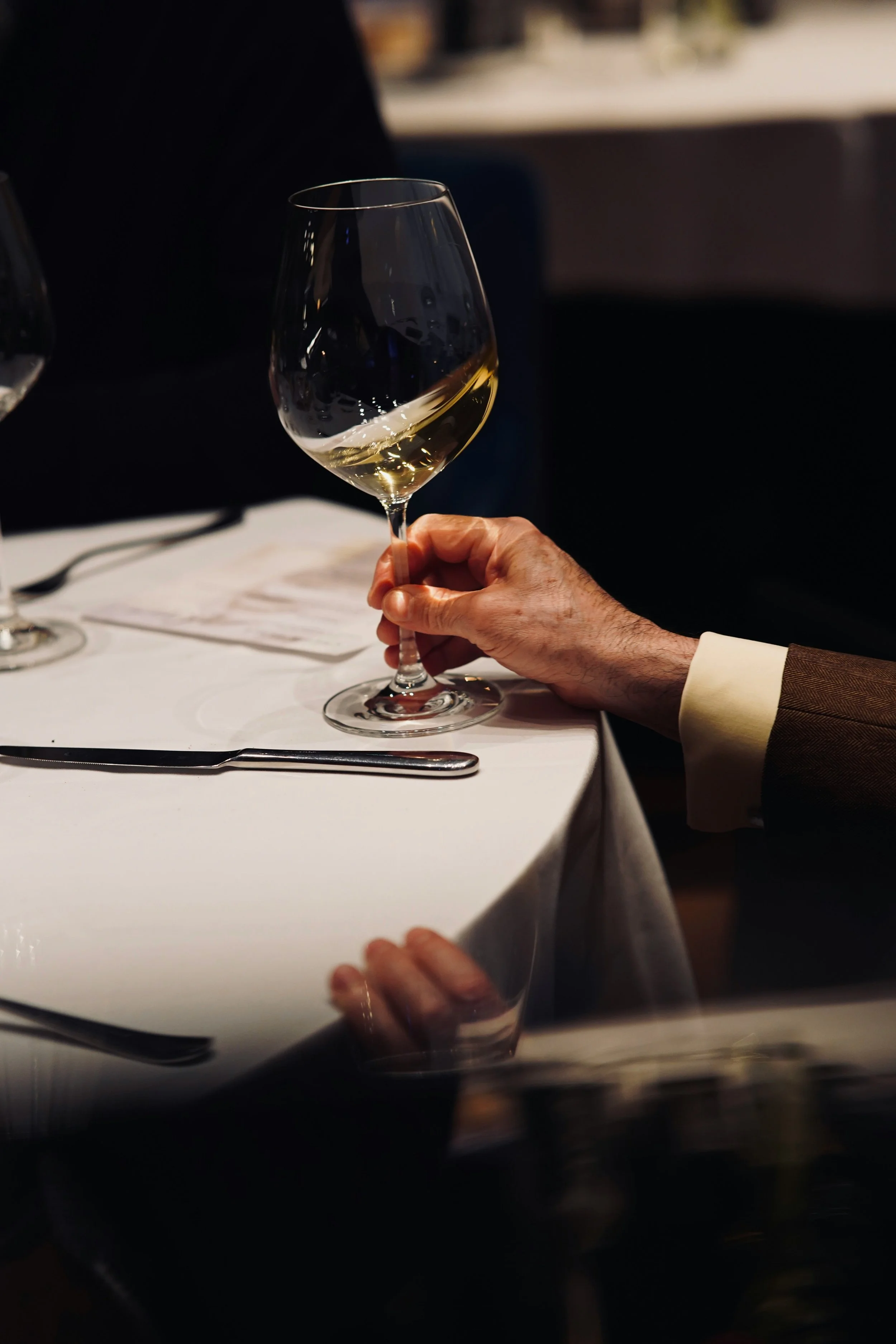 Una mano tiene in mano un bicchiere di vino bianco, su un tavolo apparecchiato con una forchetta e un coltello.