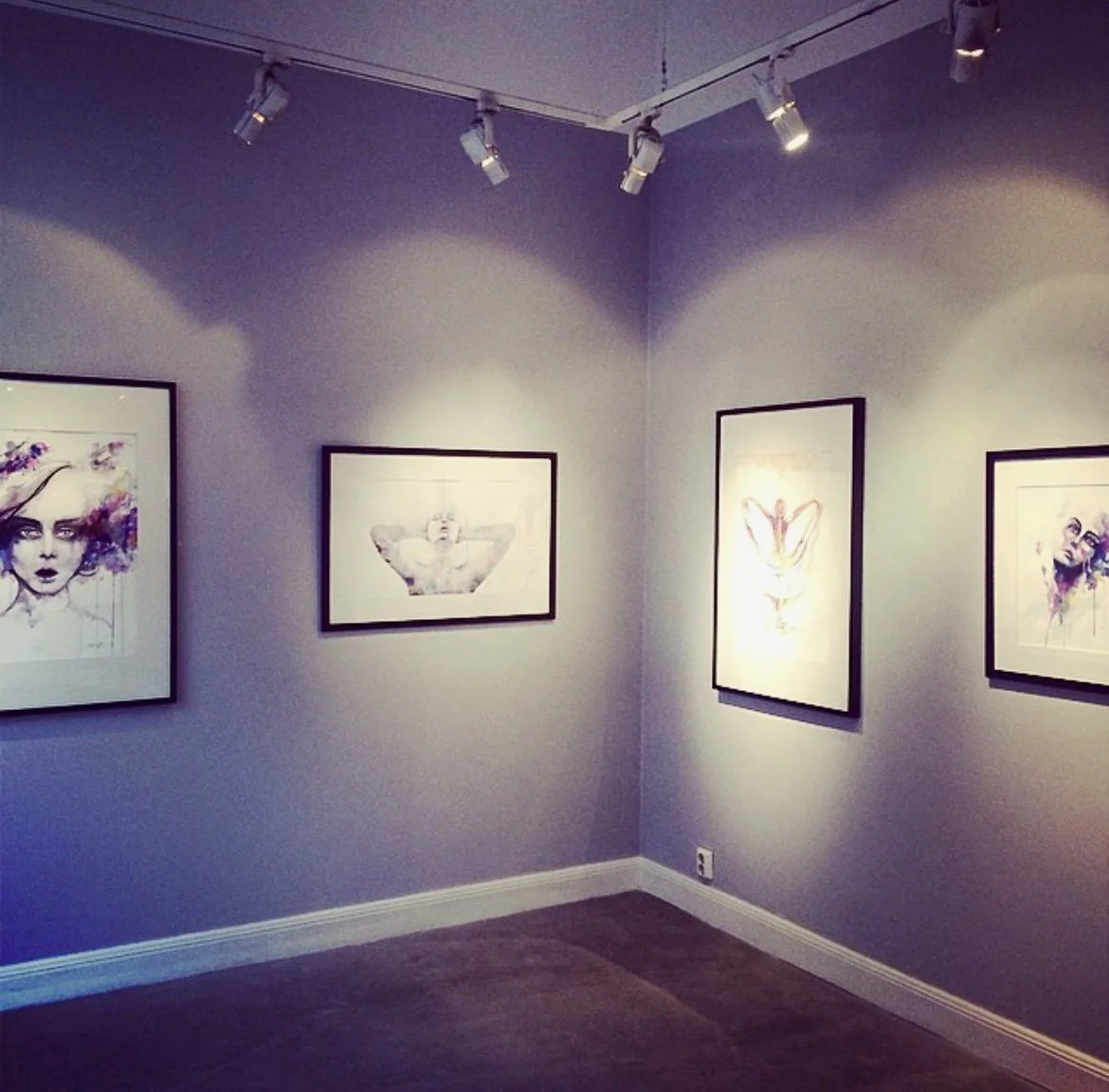 Vägg i ett konstgalleri med fyra målningar, tre av dem är porträtt av kvinnor med konstnärlig stil, ovanpå sitter spotlights för att belysa konstverket. Abante Galleri, Stockholm.