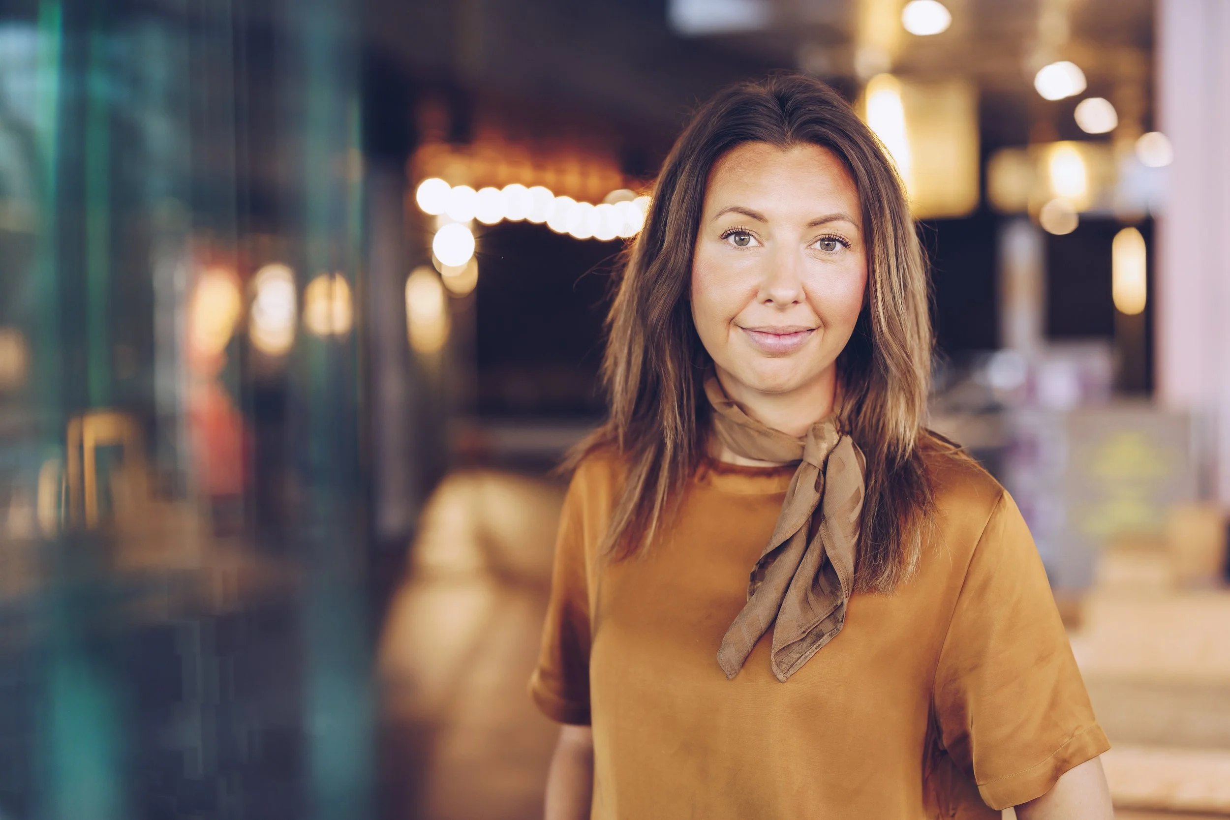 Isabelle Malmvall. Brunt hår, klädd i en orange outfit, står i en stadsmiljö med bokeh-effekt av stadens ljus i bakgrunden.
