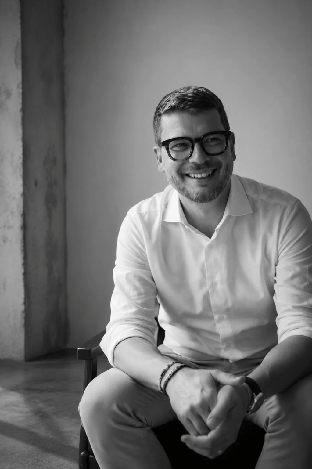 A smiling man with glasses wearing a white shirt, sitting on a chair in a room with a concrete wall and a plain background.