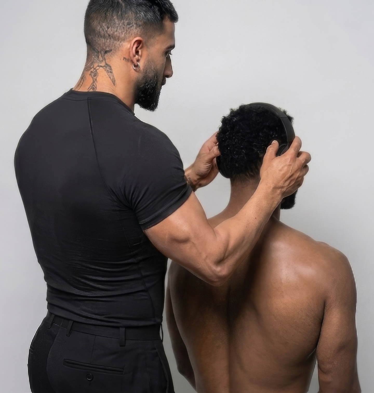 A man with tattoos on his neck and arm is holding headphones over a shirtless man's head, adjusting the headphones.