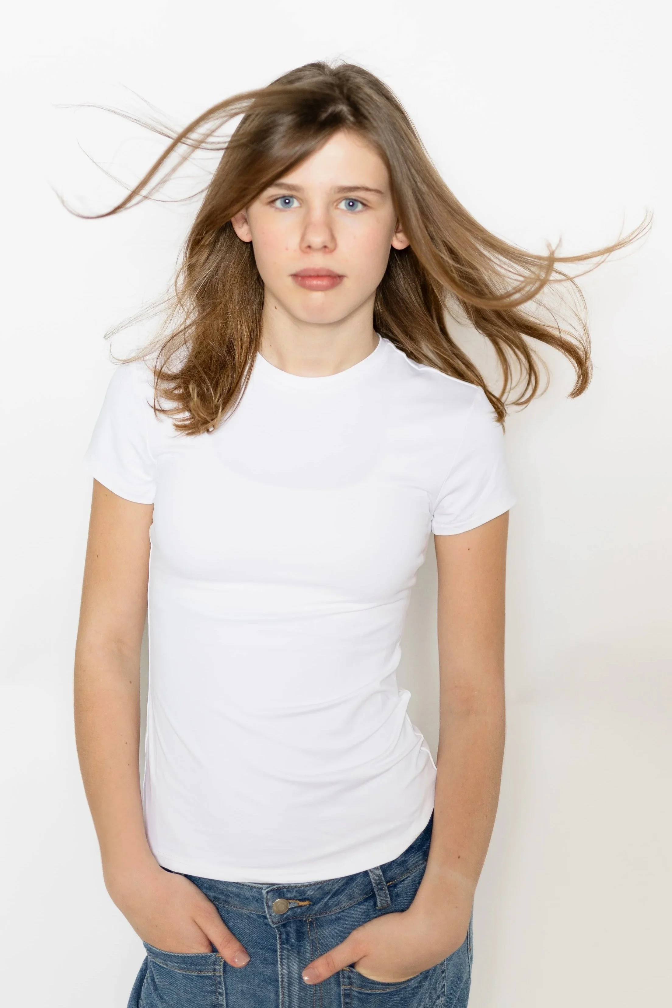 A young woman with light skin and blue eyes wearing a white t-shirt and blue jeans, standing against a plain white background, with her hands in her pockets.