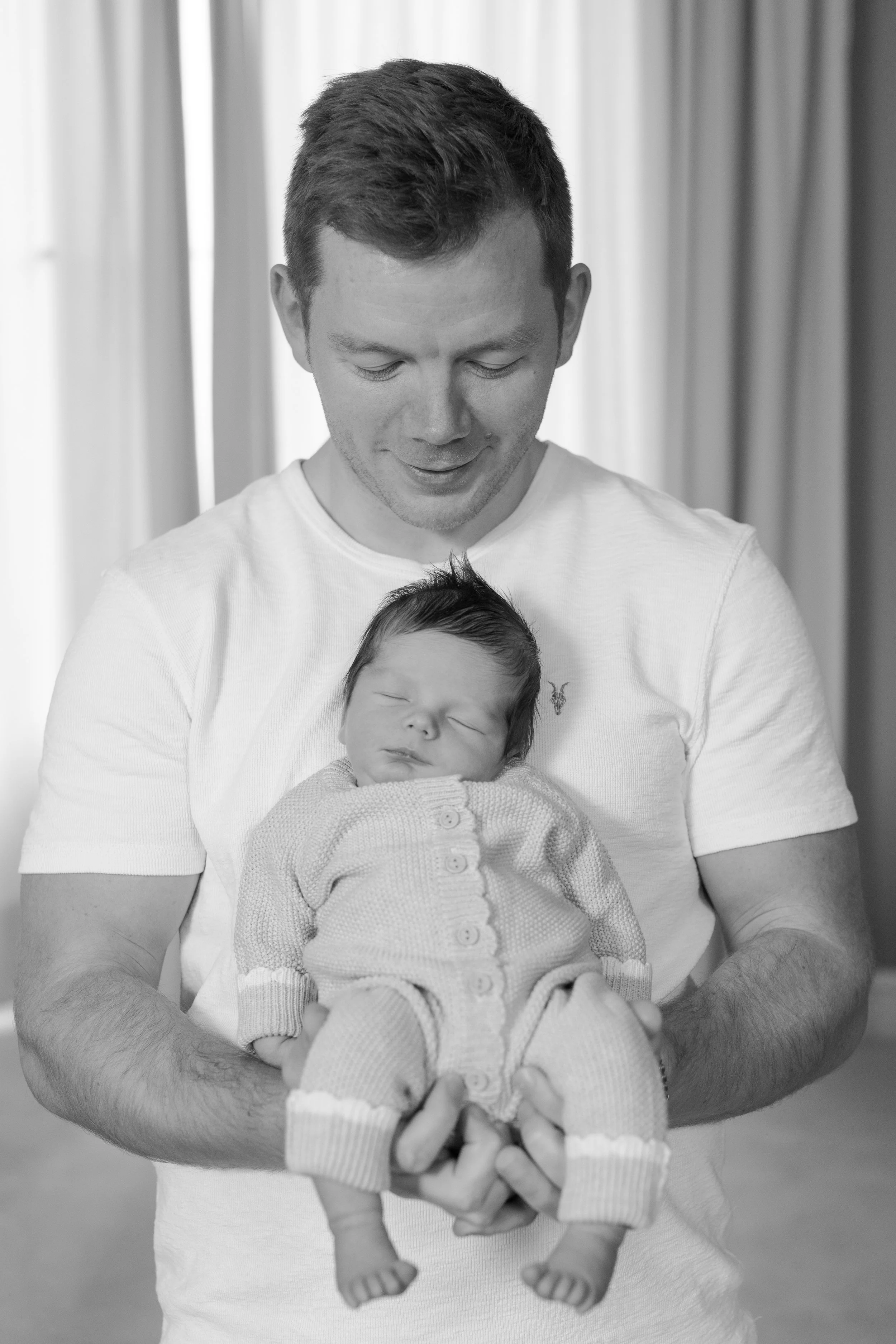 A man holding a sleeping baby, with both facing downward. The man is looking down at the baby with a gentle expression, and the baby is wrapped in a cozy sweater.