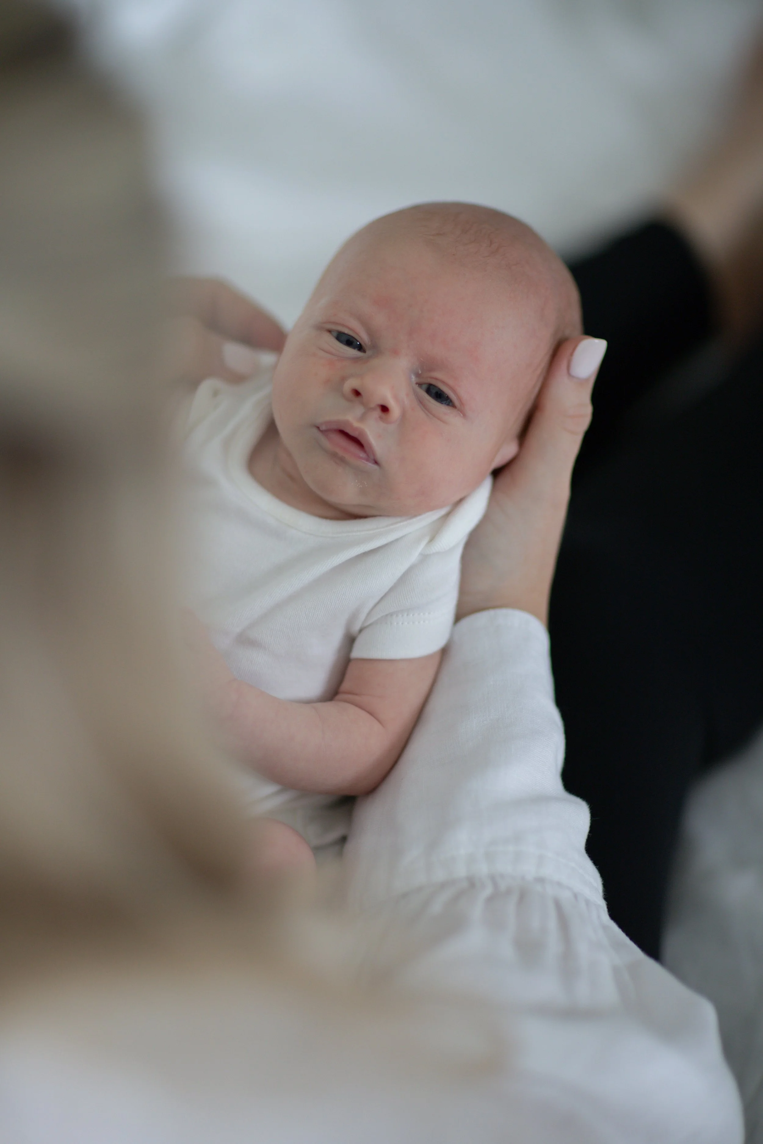 A close-up of a newborn baby with blue eyes being gently held by an adult with a hand supporting the baby's head, wearing a white outfit.