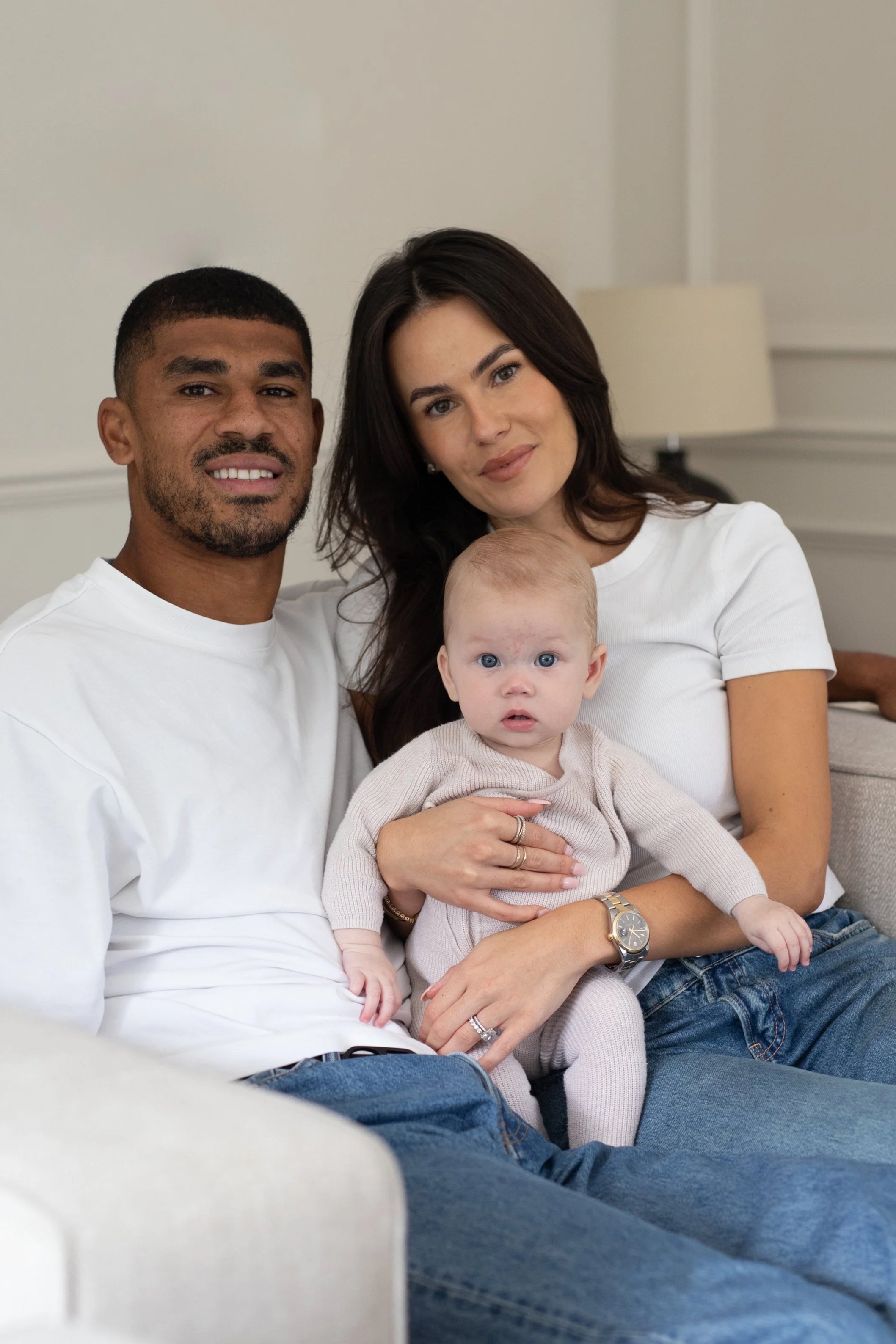 A family of three sitting on a couch in a cozy living room, with a man, woman, and a baby girl with blue eyes, all looking at the camera.