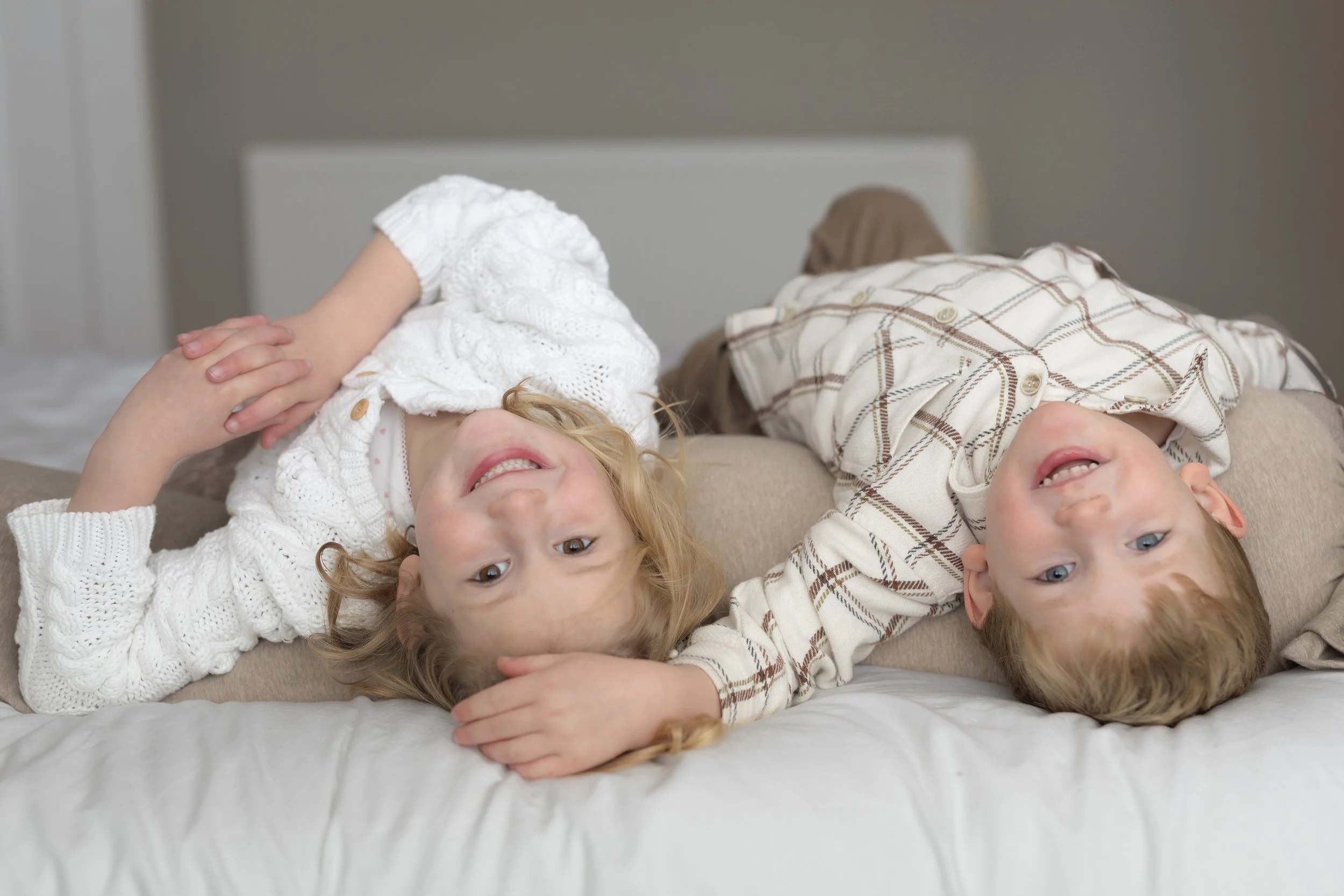 Two young children lying upside down on a bed, smiling and looking at the camera. The girl has curly blonde hair and is wearing a white sweater, while the boy has straight blonde hair and is wearing a plaid shirt.